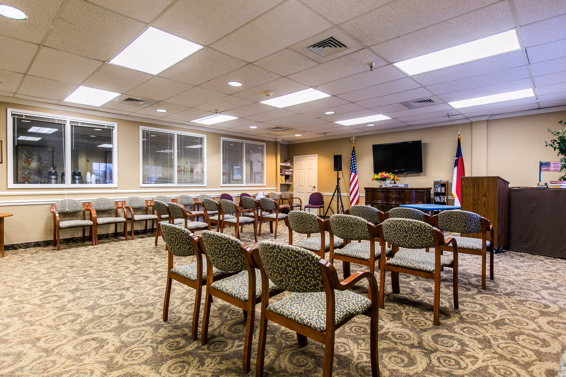 A meeting or activity room with multiple rows of patterned chairs arranged facing a podium and a flat-screen TV mounted on the wall. The room has beige walls, a patterned carpet, fluorescent ceiling lights, and two flags displayed near the podium. There are windows on one side showing another room with coffee dispensers.