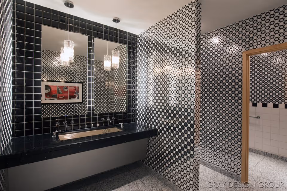 Modern bathroom with black and white circular patterned tiles on the walls, a black countertop with an integrated sink, two rectangular mirrors above the sink, and two hanging pendant lights. The floor is covered with light gray tiles, and a wooden door frame leads to another tiled area.