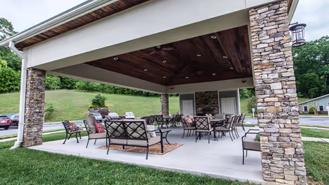 Outdoor covered seating area with stone pillars and wooden ceiling, furnished with multiple chairs and tables on a concrete floor, surrounded by grass and trees in the background.