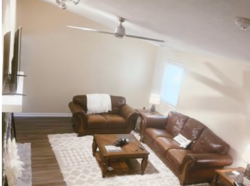 A cozy living room with two brown leather sofas, a wooden coffee table, a white patterned rug, a ceiling fan, and a window with blinds. There is a lamp on a side table next to one of the sofas and a TV mounted on the wall.