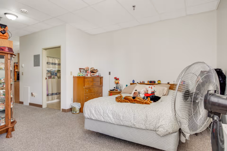 A bedroom with a bed covered in a light-colored patterned comforter and several stuffed animals on top. To the right of the bed is a large standing fan. On the left side of the room is a wooden dresser with various items on top, and an open door leading to a bathroom with a shower curtain visible. The room has beige walls and carpeted floor.