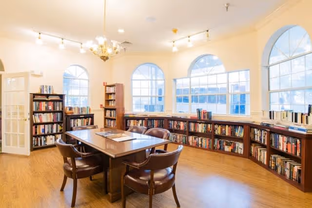 A bright and spacious library room with large arched windows letting in natural light. The room features wooden bookshelves filled with books along the walls and a central wooden table surrounded by six brown leather chairs. The floor is wooden, and there is a chandelier and ceiling lights providing additional illumination.
