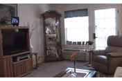 A cozy living room with a large flat-screen TV on a wooden stand, a tall glass display cabinet with decorative items, a comfortable recliner chair, a small table with a flower vase, and two windows with curtains allowing natural light to enter.