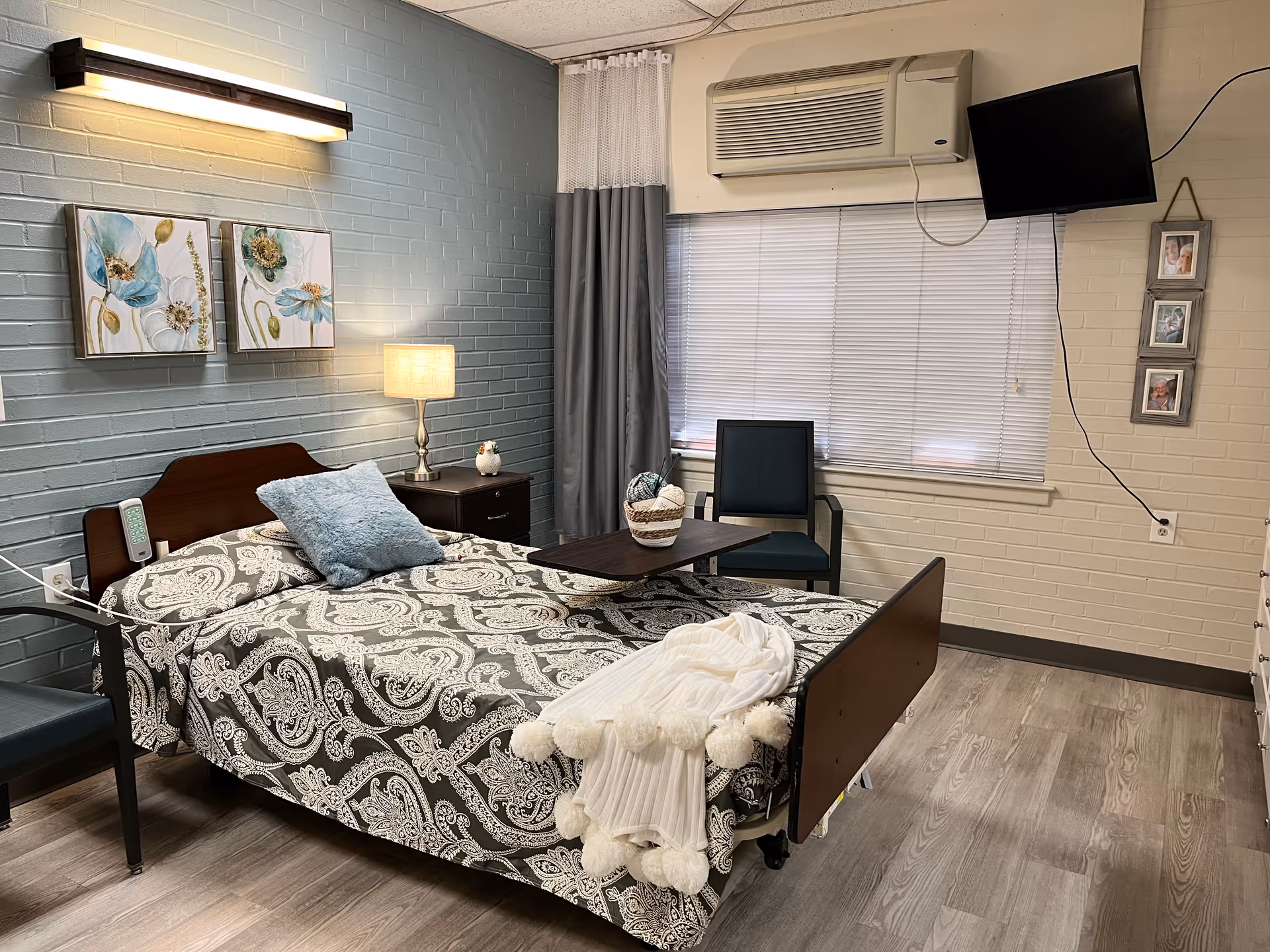 A neatly arranged nursing facility bedroom with a single bed covered in a patterned bedspread and a light blue pillow. There is a small bedside table with a lamp and decorative item, a chair, a wall-mounted TV, and window blinds. The walls are painted in light gray and beige tones with two floral paintings above the bed.