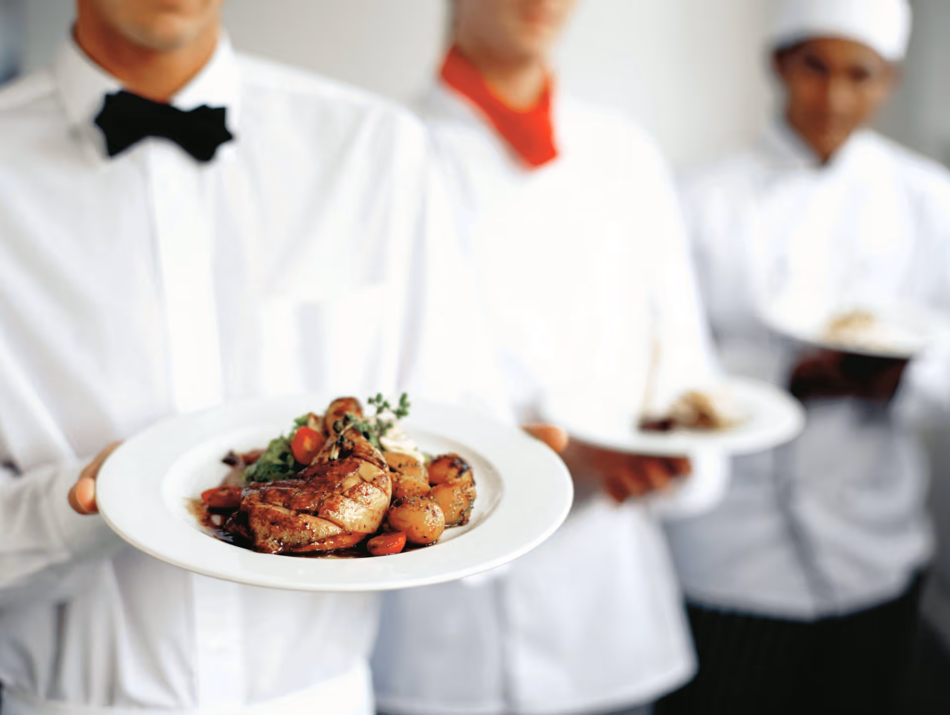 Three servers dressed in white uniforms holding plates of gourmet food, with the focus on the front plate featuring a cooked chicken leg, potatoes, and vegetables.