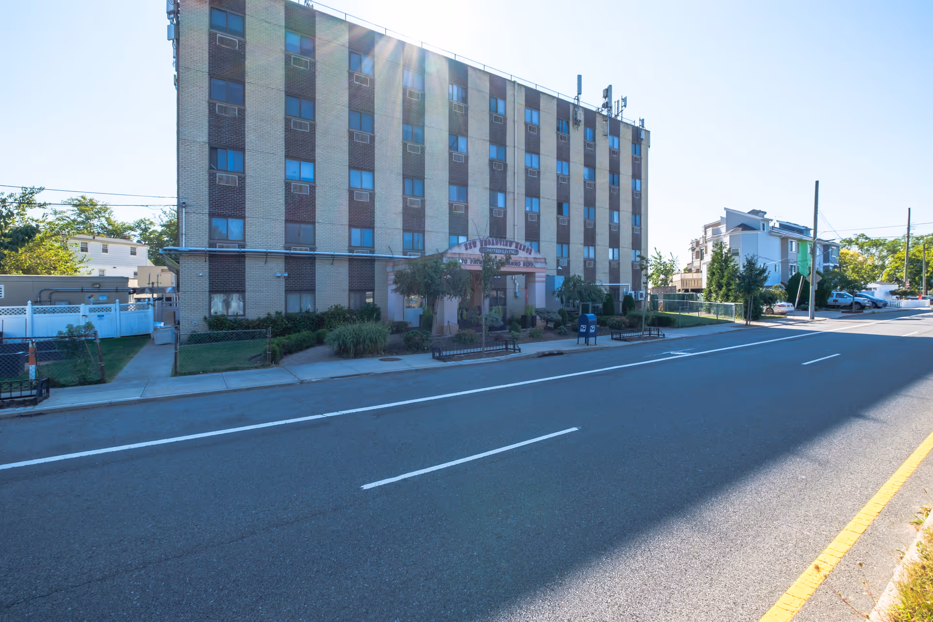 Five-story brick residential building with an awninged entrance, landscaping, and a roadway in front.