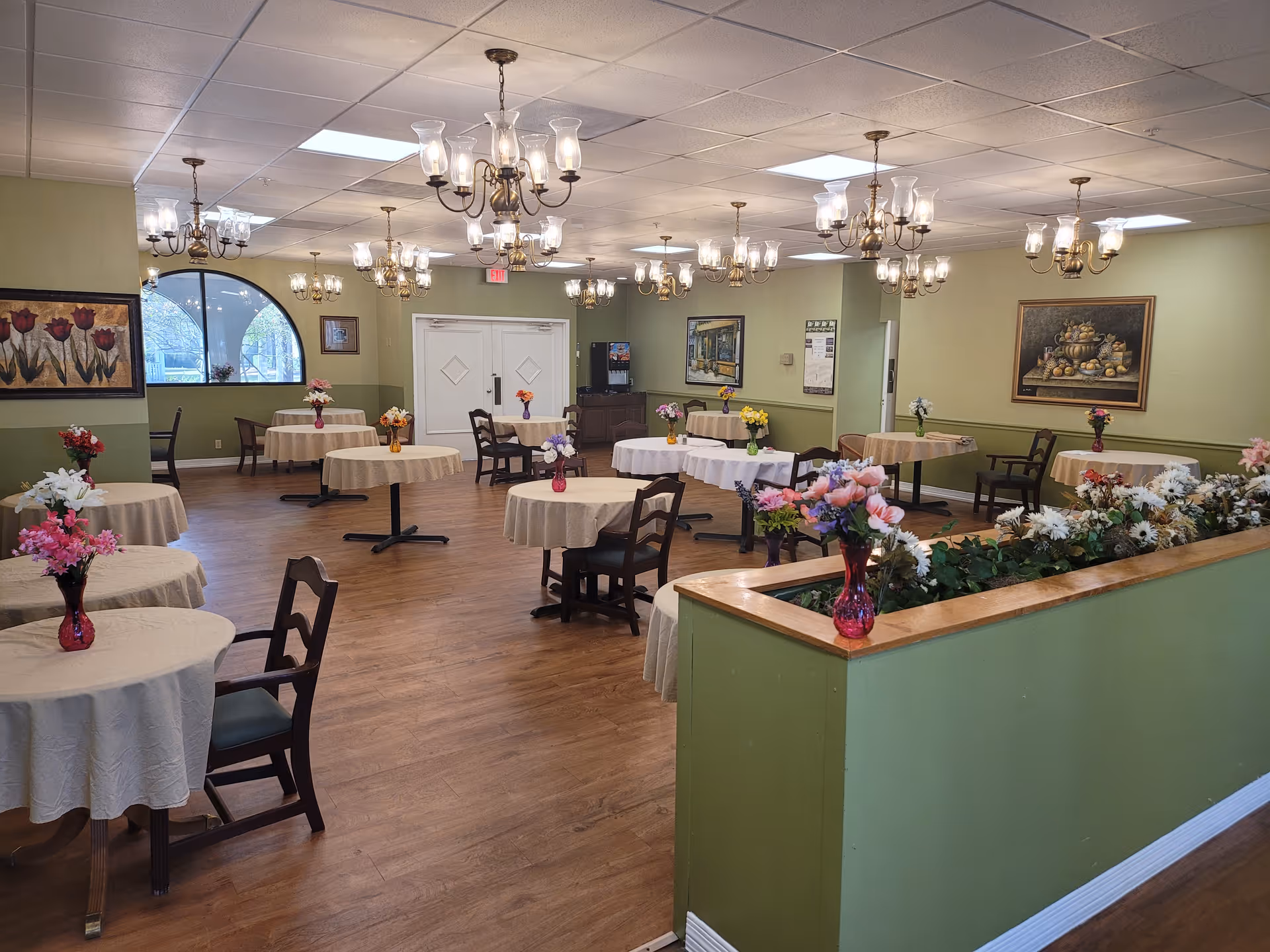 A spacious dining room with multiple round tables covered with beige tablecloths, each adorned with a vase of colorful flowers. The room has wooden flooring, green walls, several chandeliers hanging from the ceiling, and framed paintings on the walls. There is a half-wall planter with white flowers in the foreground and a large window on the left side.