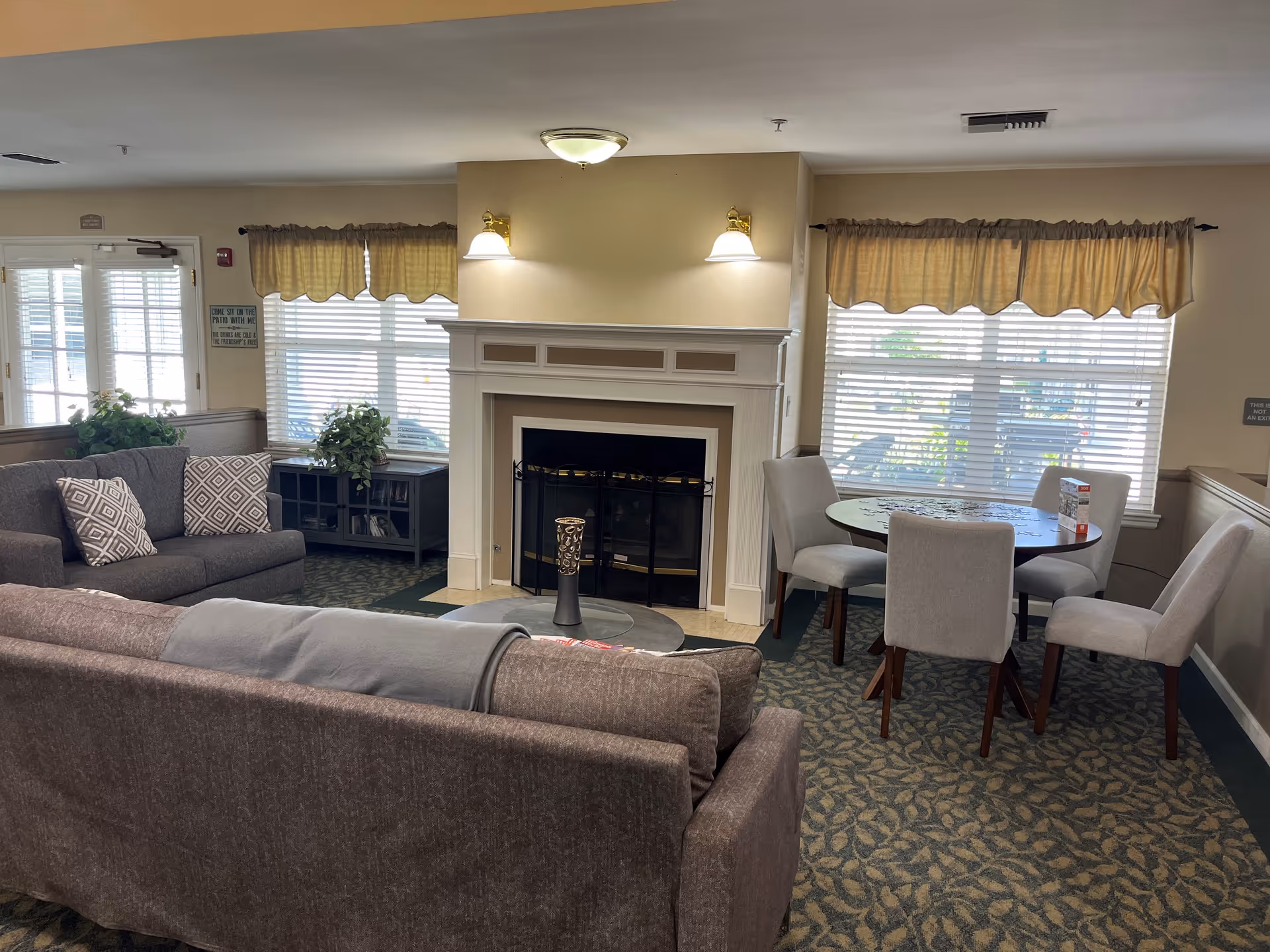 A cozy living room area with two gray sofas, a round table with four chairs, a fireplace in the center, and windows with beige curtains letting in natural light. There are plants on a small cabinet near the window and patterned carpet on the floor.