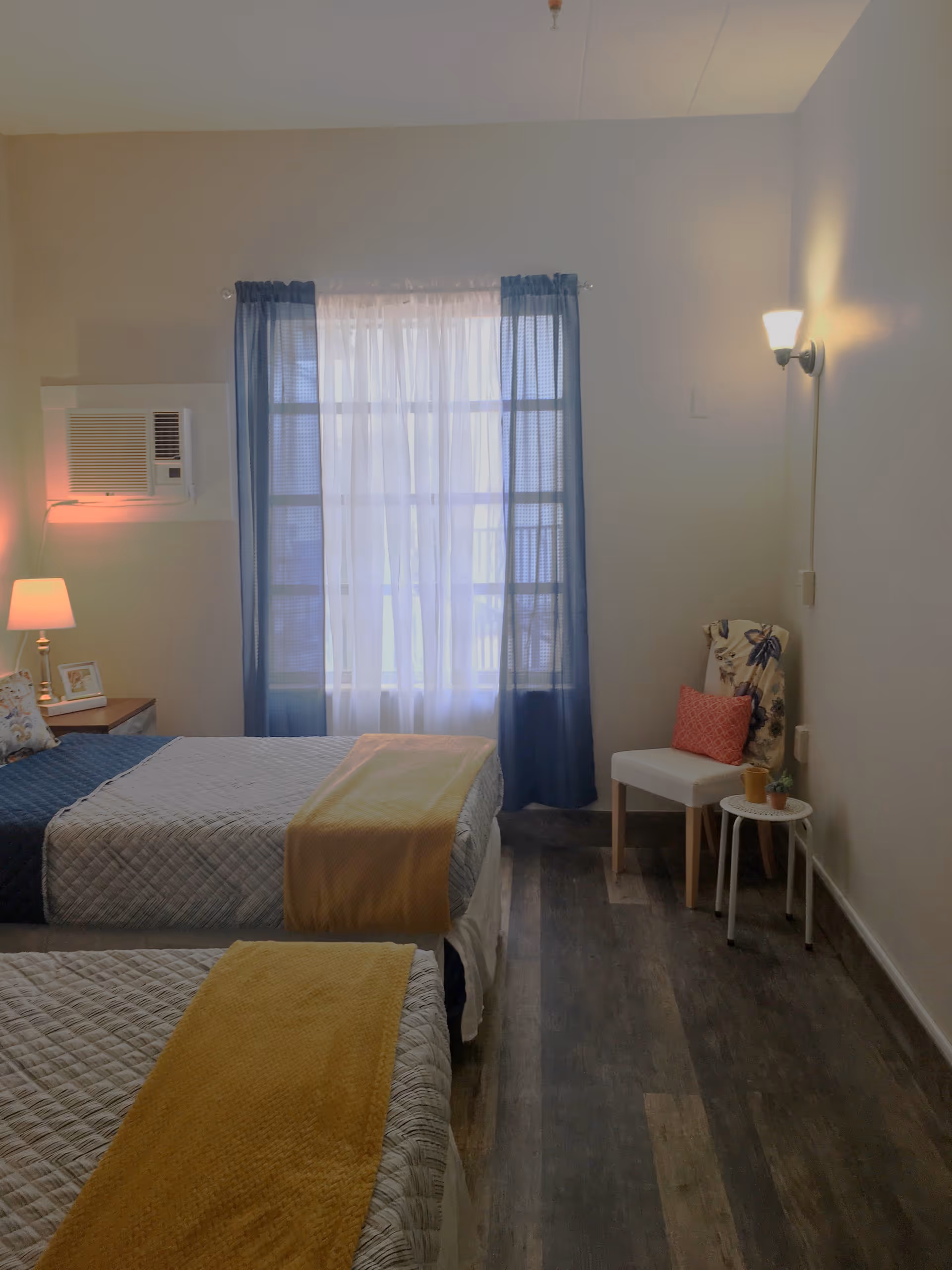 A cozy bedroom with two twin beds, each covered with a quilted bedspread and a yellow throw blanket. A window with sheer white curtains and blue drapes is centered on the far wall. To the right, there is a white chair with a floral cushion and a red pillow, next to a small round white table holding a small plant and a cup. A wall-mounted light fixture is above the chair, and a bedside table with a lamp and picture frame is visible beside one of the beds. The floor is wood with a dark finish.