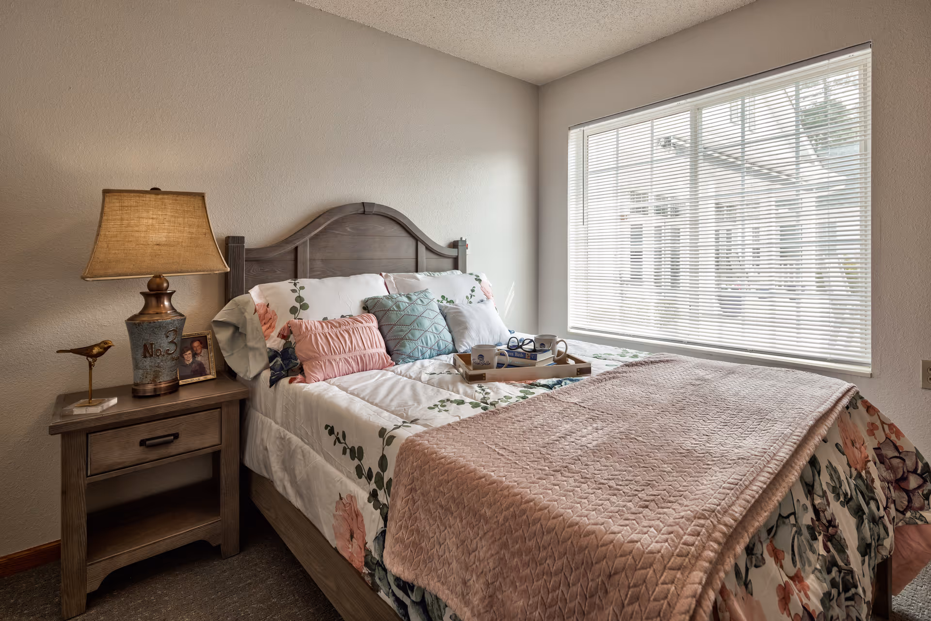 A cozy bedroom with a wooden bed frame and floral bedding, featuring multiple pillows and a pink textured blanket. A wooden nightstand beside the bed holds a lamp, a small bird figurine, and a framed photo. A large window with blinds allows natural light to fill the room.