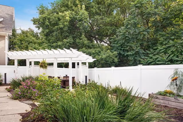 Outdoor garden area with a white pergola, surrounded by green plants and flowers, a white fence, and trees in the background.