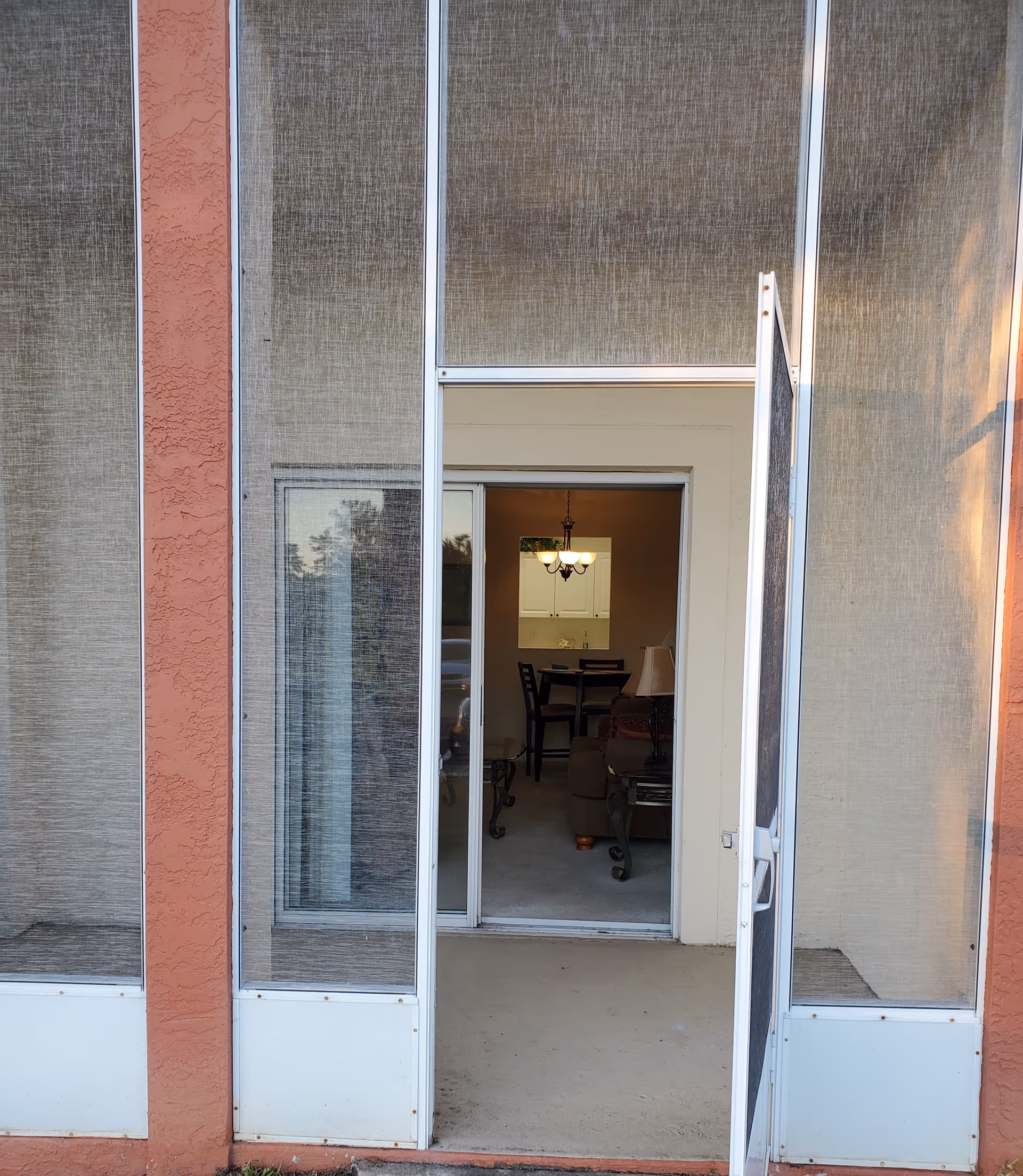 View through a screened patio door into a living room area with a sofa, a side table with a lamp, and a dining area with a table and chairs under a chandelier light fixture. The patio has a concrete floor and is enclosed with mesh screens framed in white.