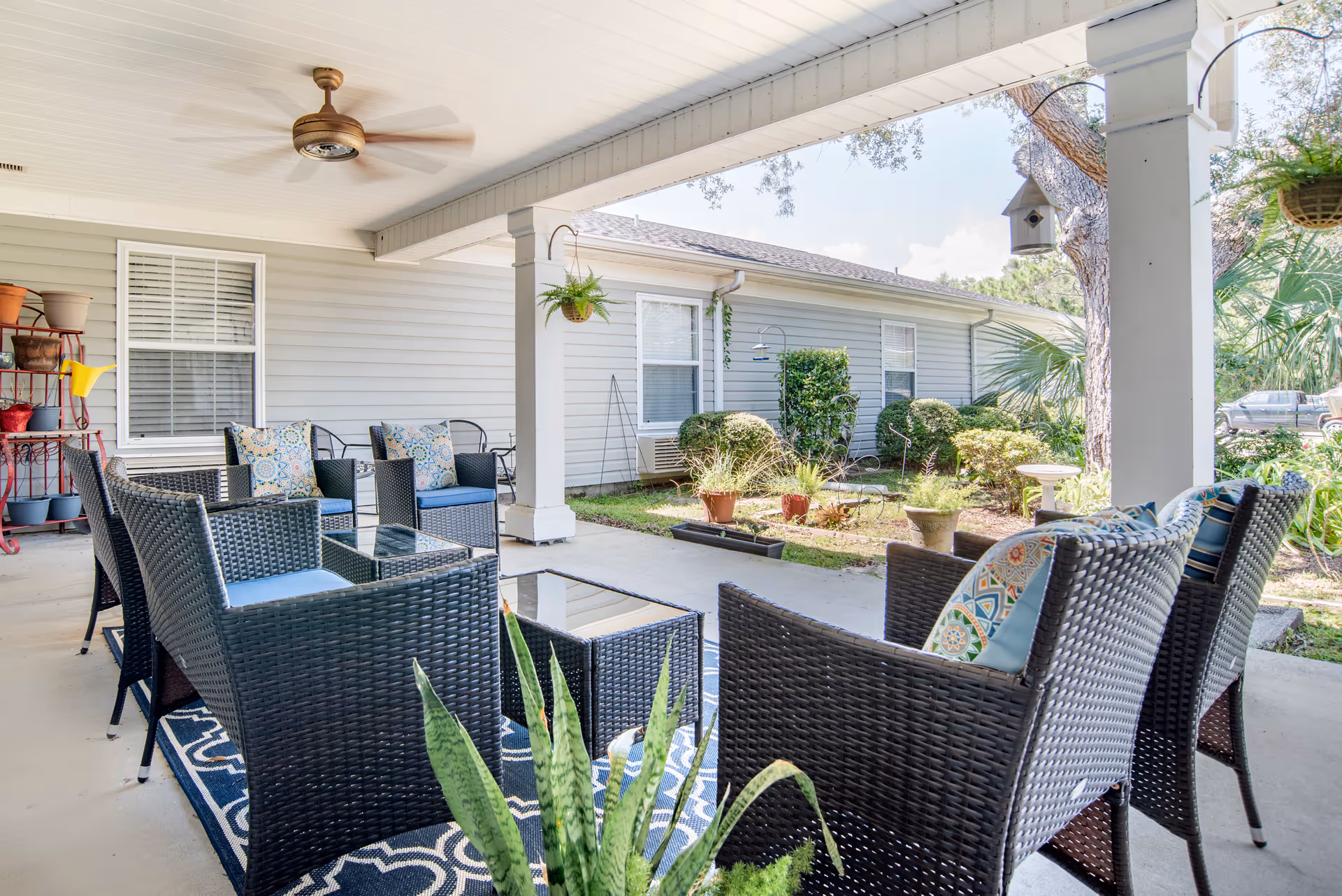 Covered outdoor patio with wicker seating, patterned cushions, a ceiling fan and potted plants facing a landscaped garden.