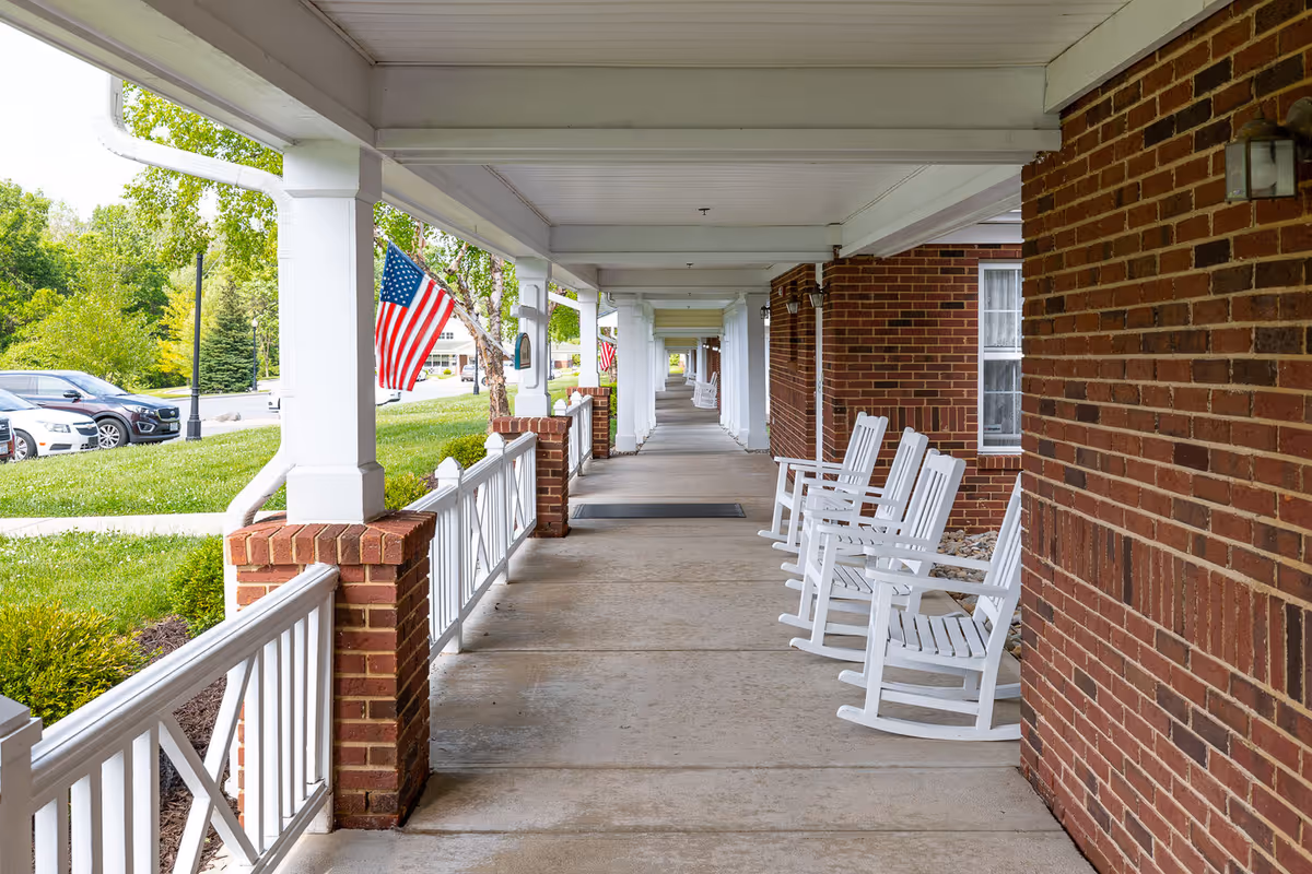 Covered outdoor walkway with white rocking chairs lined up along a brick wall. White columns support the roof, and American flags are displayed on the left side near a grassy area and parked cars.
