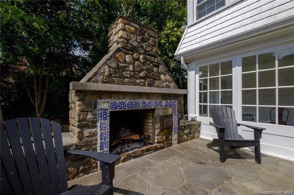 Outdoor stone fireplace with blue and white decorative tiles around the firebox, flanked by two black Adirondack chairs on a stone patio next to a white building with large windows.