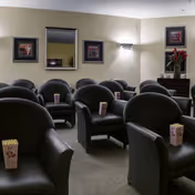 A small screening room with rows of black armchairs, each holding a popcorn box, mirrored and framed artwork on the wall, and a side table with flowers.
