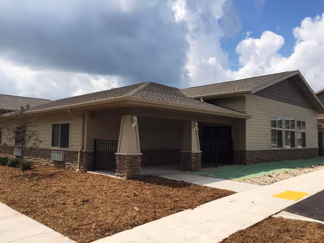 Single-story building exterior with a covered entrance supported by stone pillars, adjacent sidewalk and landscaped beds under a partly cloudy sky.