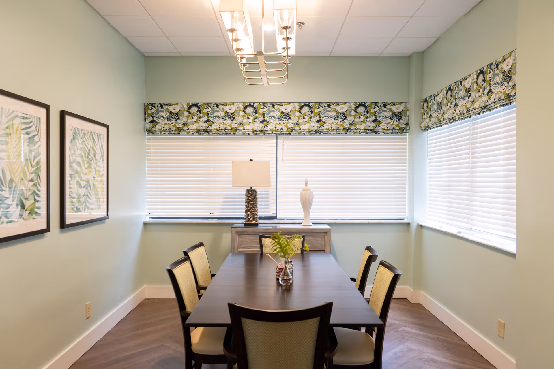 A dining room with a dark wooden table surrounded by six chairs with light-colored upholstery. The room has light green walls, two framed botanical prints on the left wall, and two windows with white blinds and floral valances. A sideboard with a lamp and a decorative vase is positioned against the back wall beneath the windows. A modern rectangular chandelier hangs from the ceiling above the table.
