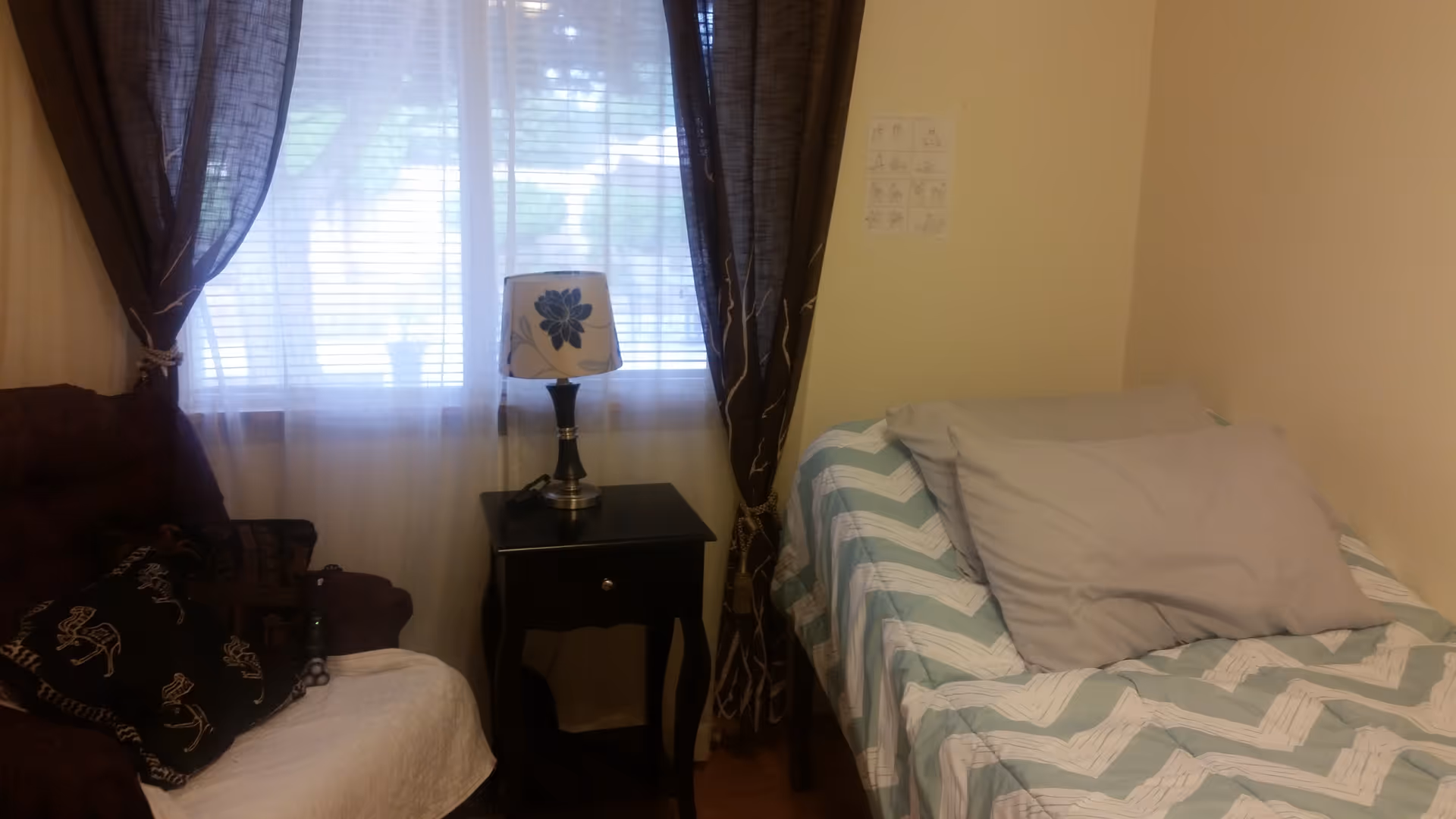 Small bedroom with a single bed covered in a chevron comforter, a nightstand and lamp beside a curtained window and an armchair.