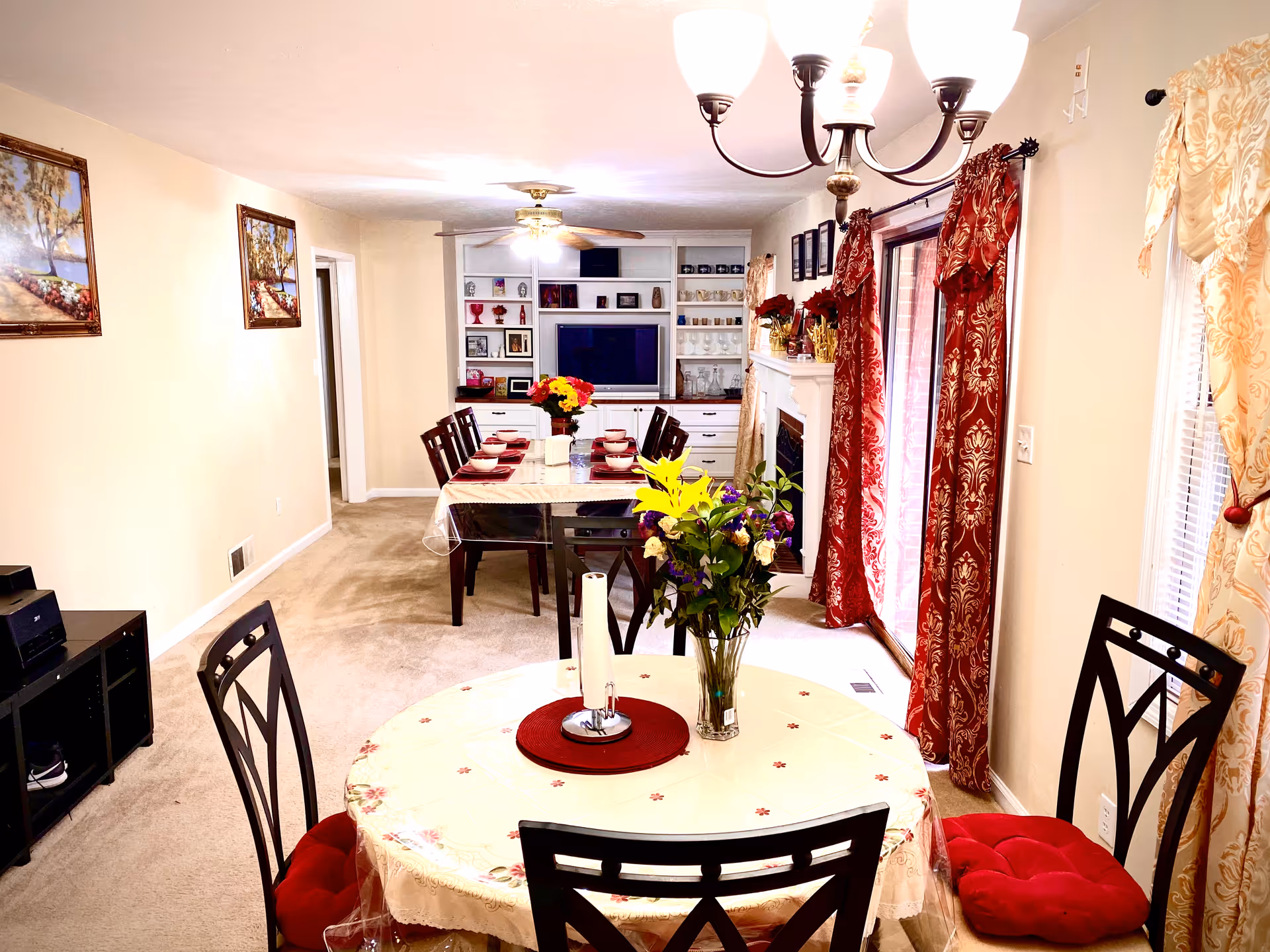 A bright and cozy dining and living area in a senior living facility. The foreground shows a round table with a floral tablecloth, a vase of flowers, and four chairs with red cushions. In the background, there is a long dining table set with bowls and plates, surrounded by chairs. The room has beige walls, carpeted floors, red and gold patterned curtains on the windows and sliding glass door, a ceiling fan, a chandelier, and a built-in white shelving unit with a TV and decorative items.