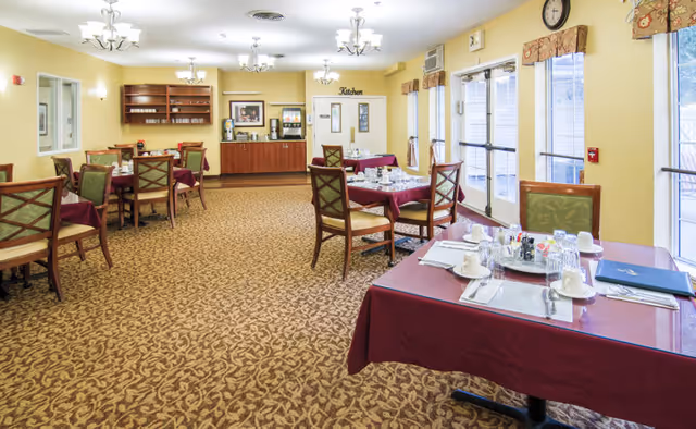 A dining room in a senior living facility with several tables covered in burgundy tablecloths, set with white cups, plates, and silverware. The room has patterned carpet, yellow walls, large windows with valances, and a serving area labeled 'Kitchen' in the background.