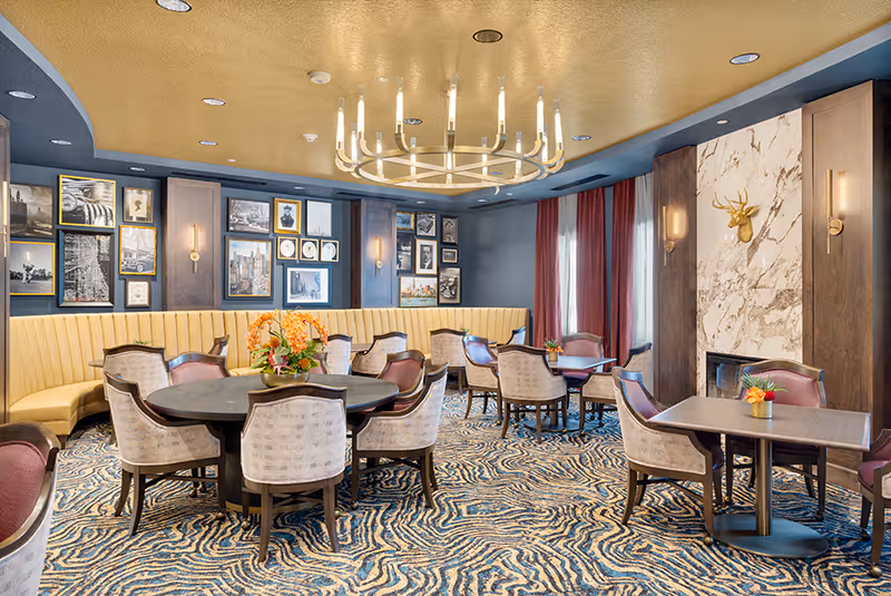 A stylish dining room with round and square tables surrounded by upholstered chairs. The room features a yellow curved banquette along one wall, a large modern chandelier hanging from a gold ceiling, patterned carpet, and a marble fireplace with a gold deer head mounted above it. The walls are decorated with framed black and white and color photographs, and there are red curtains on the windows.