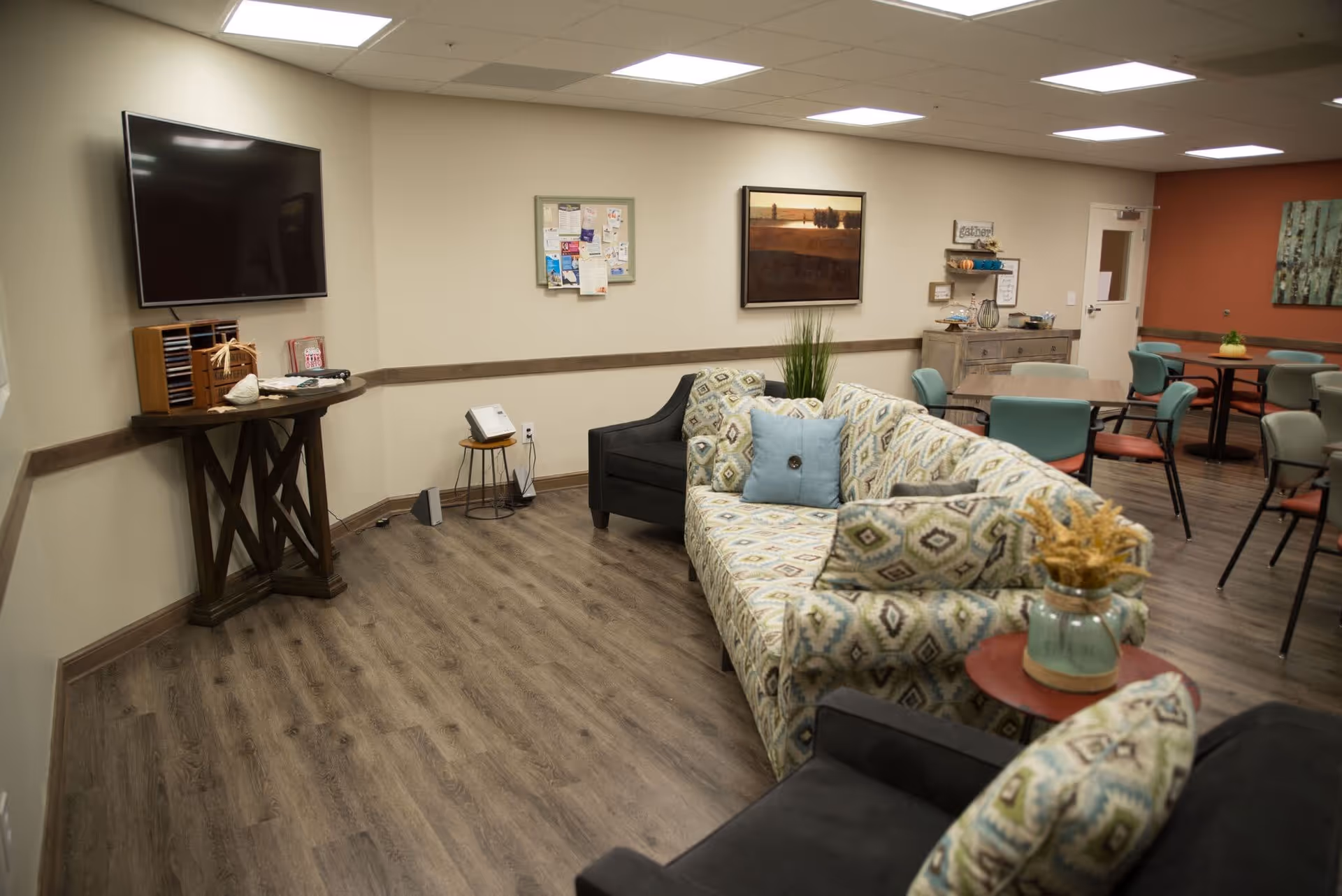 A cozy common area with patterned sofas and a black armchair arranged around a small red side table with a decorative vase. The room has wood flooring, a wall-mounted flat-screen TV, a bulletin board, and framed artwork on the walls. In the background, there are several tables with chairs, a wooden cabinet with decorative items, and a door.