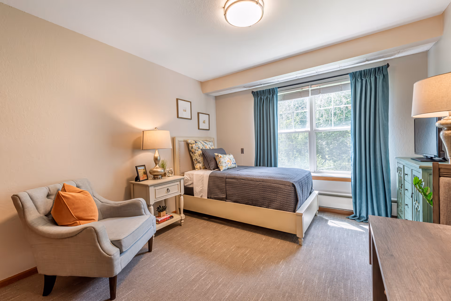 A cozy bedroom with a single bed dressed in gray bedding and floral pillows, a beige armchair with an orange cushion, a nightstand with a lamp, clock, and small plant, a window with blue curtains letting in natural light, and a green cabinet with a TV on top.