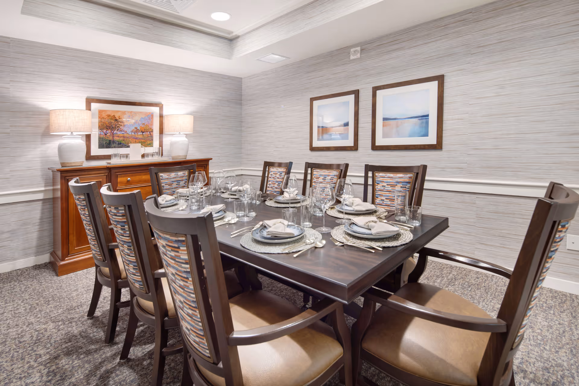 A formal dining room with a dark wooden table set for eight people. Each place setting includes a plate, napkin, glassware, and silverware. The chairs have wooden frames with patterned fabric backs and cushioned seats. Against the wall is a wooden sideboard with two lamps and a framed painting above it. The walls are covered with light-colored textured wallpaper and two framed landscape pictures hang on the adjacent wall.