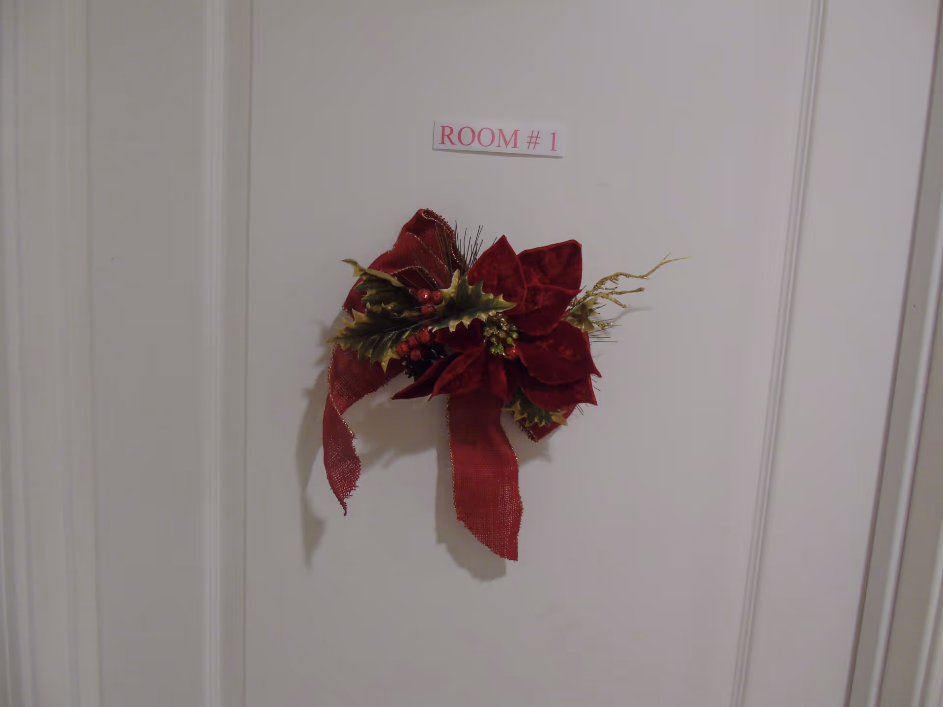 A white door with a small sign above it that reads 'ROOM # 1' and a decorative red poinsettia flower with green leaves and a red ribbon attached to the door.
