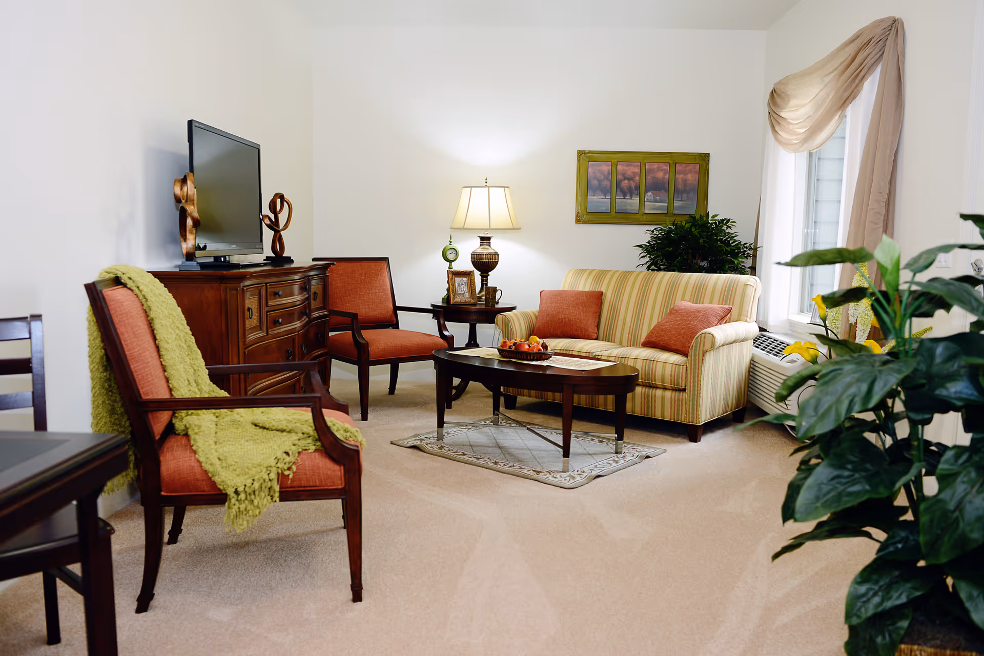 A cozy living room with a striped sofa with two orange cushions, two orange armchairs, a wooden coffee table with a fruit basket, a wooden TV stand with a flat-screen TV and decorative sculptures, a side table with a lamp, picture frame, and clock, a window with beige curtains, and several green plants.