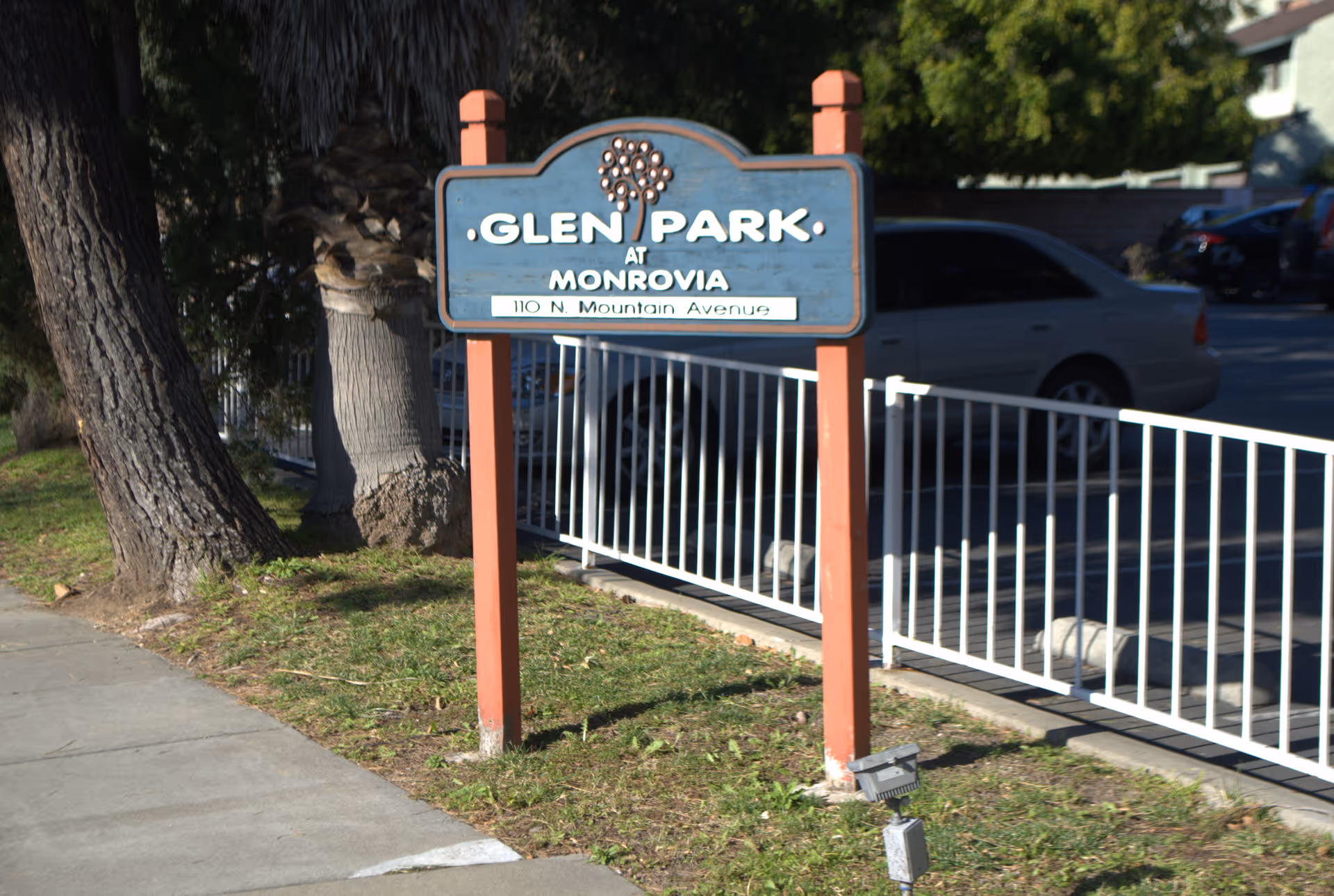A wooden sign reading "Glen Park at Monrovia" on red posts beside a sidewalk and white metal fence with parked cars behind.