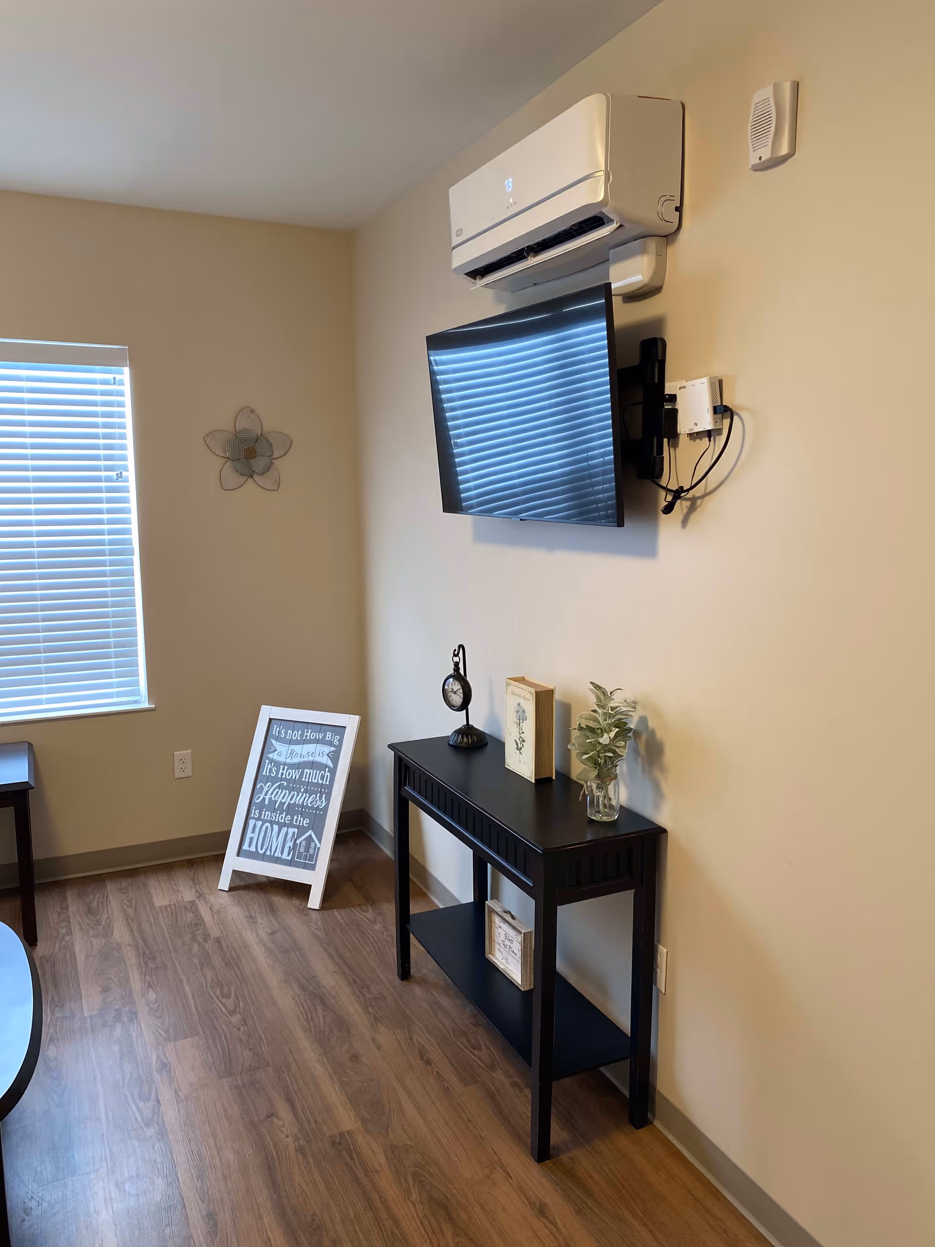 A corner of a room with a mounted flat-screen TV below a wall-mounted air conditioning unit. Below the TV is a black console table with decorative items including a small clock, a book, and a small plant in a glass jar. To the left, there is a window with closed blinds and a framed sign on the floor that reads 'It's not How Big a House is It's How much Happiness is inside the HOME'. The room has light-colored walls and wood flooring.