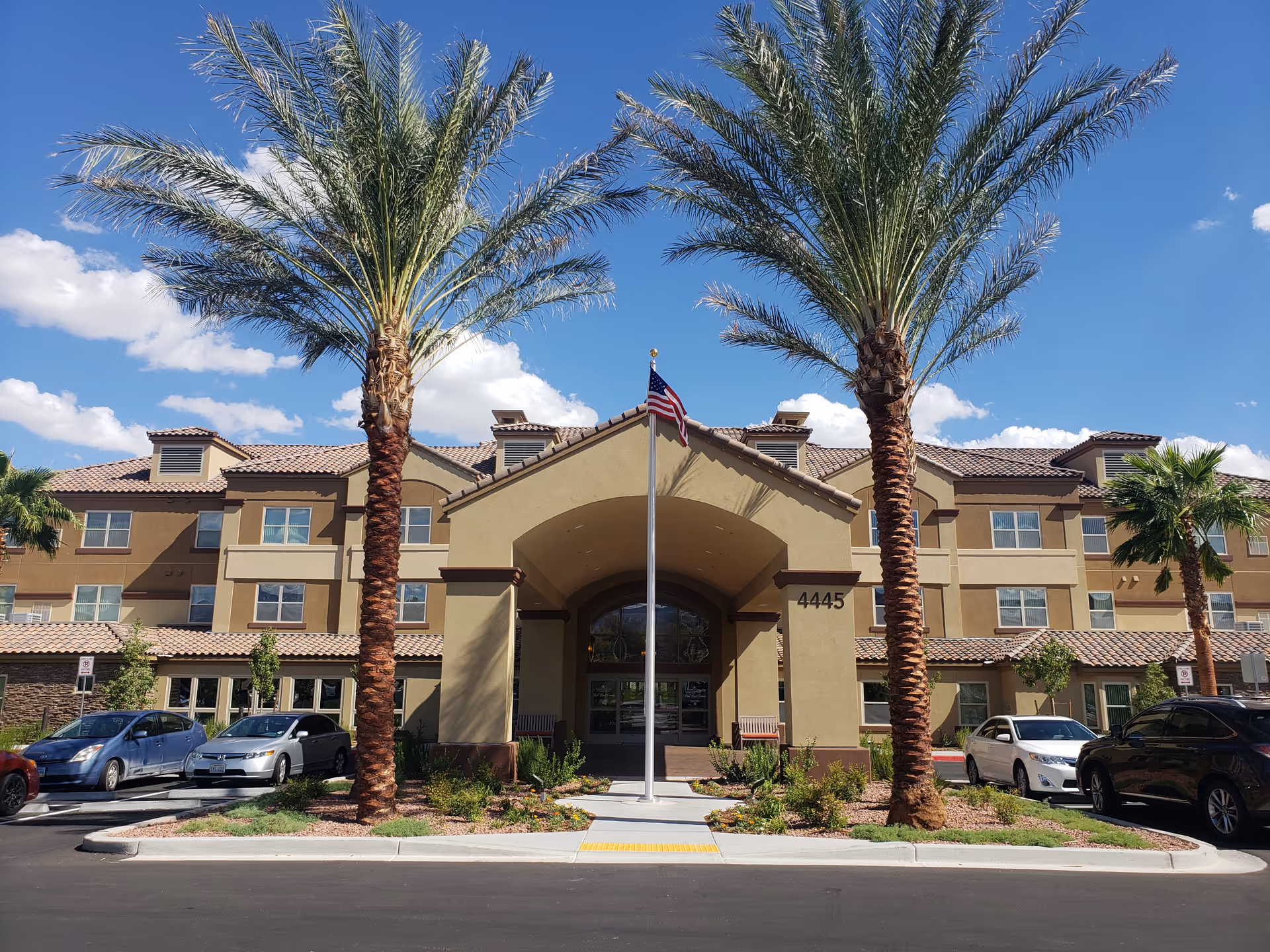 Front entrance of a three-story retirement community building flanked by palm trees and an American flag in front.