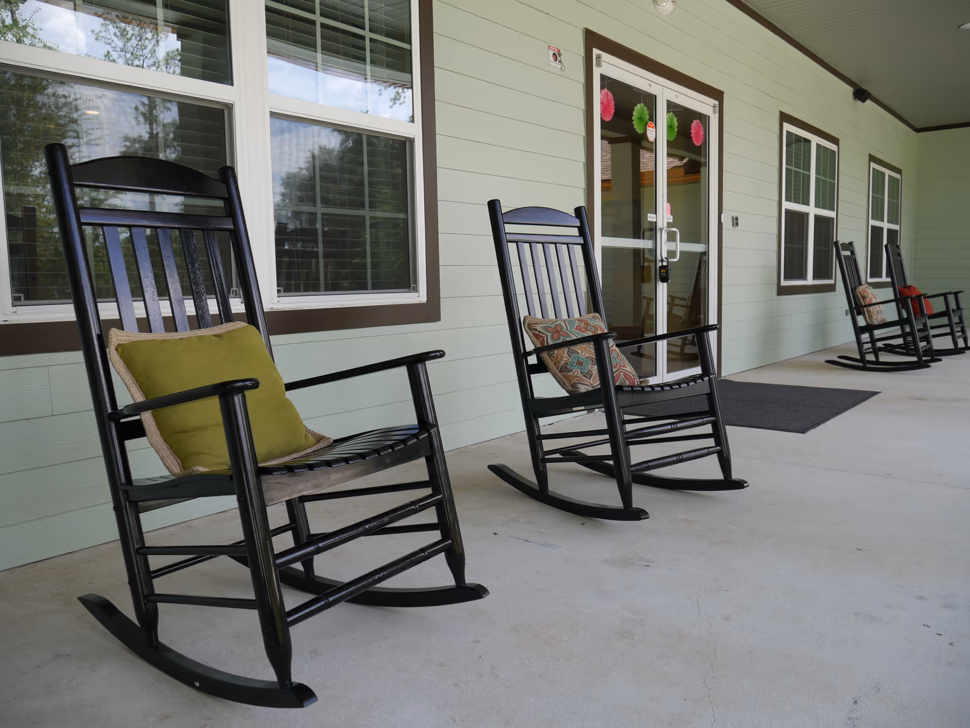 A covered porch area with several black wooden rocking chairs, each with a decorative cushion, placed along the wall of a light green building with multiple windows and a glass door.