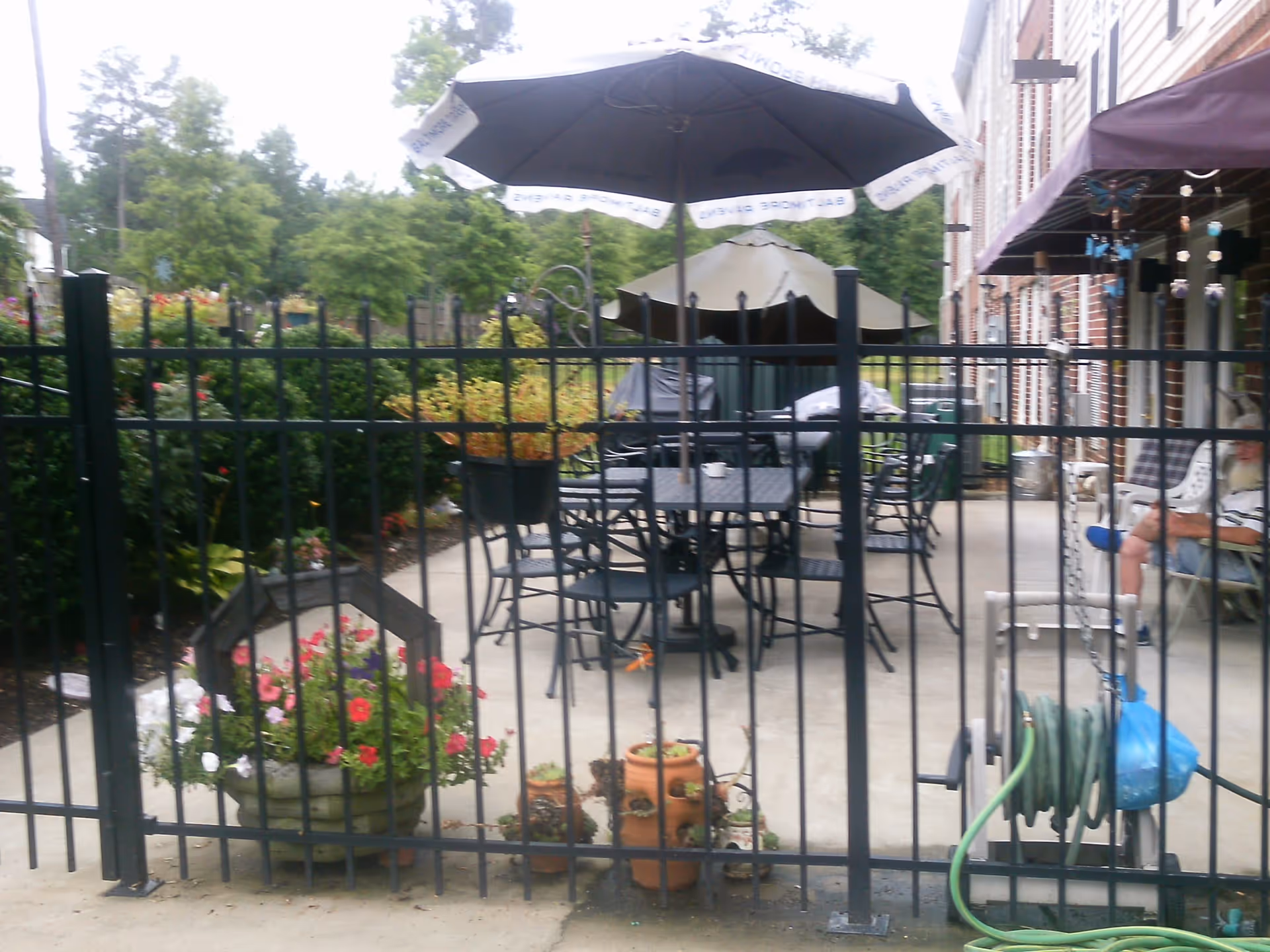 Fenced outdoor patio with tables, chairs, umbrellas and potted plants beside a brick building.