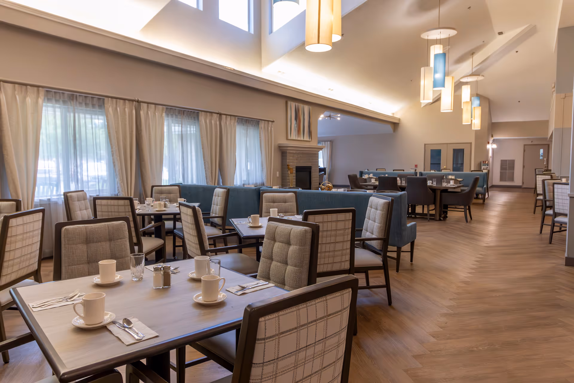 A spacious dining room with multiple tables and cushioned chairs arranged neatly. Each table is set with white cups, saucers, glasses, and silverware. Large windows with sheer curtains allow natural light to fill the room. The ceiling features modern pendant lights, and there is a fireplace with artwork above it in the background.
