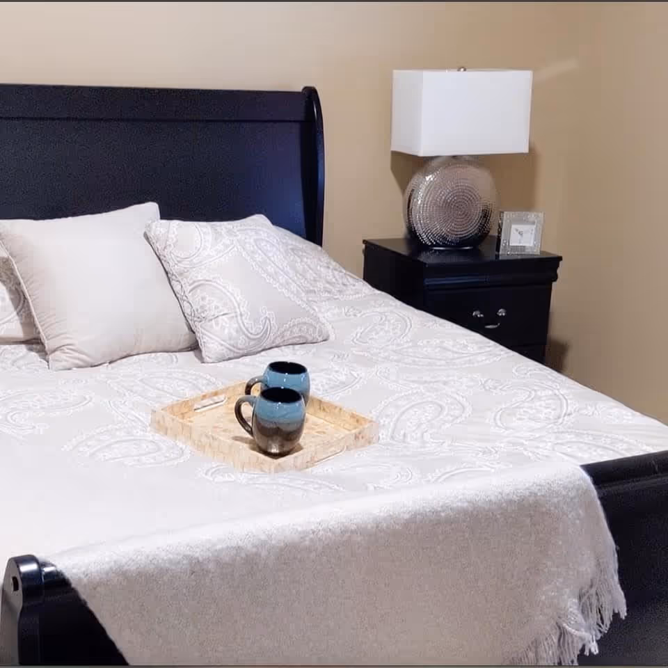 A neatly made bed with a dark wooden headboard and footboard, covered with a white patterned bedspread and two decorative pillows. On the bed is a wooden tray holding two blue and black mugs. Next to the bed is a black nightstand with a decorative lamp and a small framed picture. The walls are painted beige.