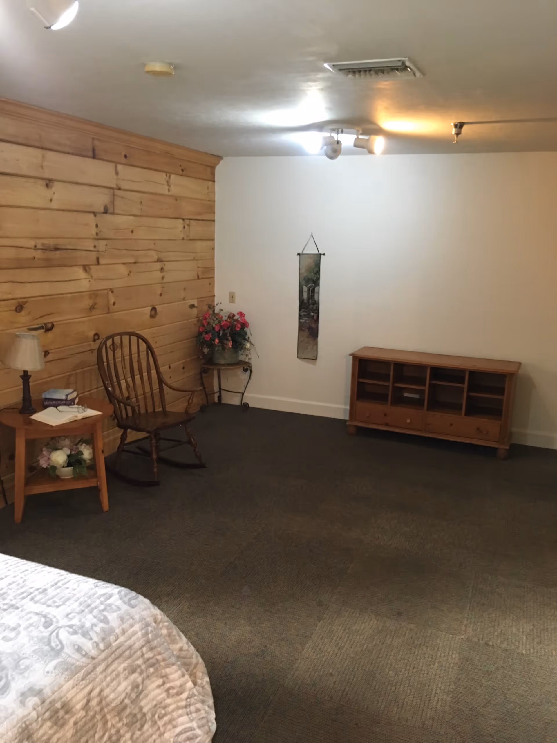 A cozy room with a wooden rocking chair, a small wooden side table with a lamp and books, a floral arrangement on a small round table, a wooden cabinet with open shelves and drawers, and a wall hanging. The room has a wood-paneled wall on one side and a plain white wall on the other, with carpeted flooring and ceiling lights.