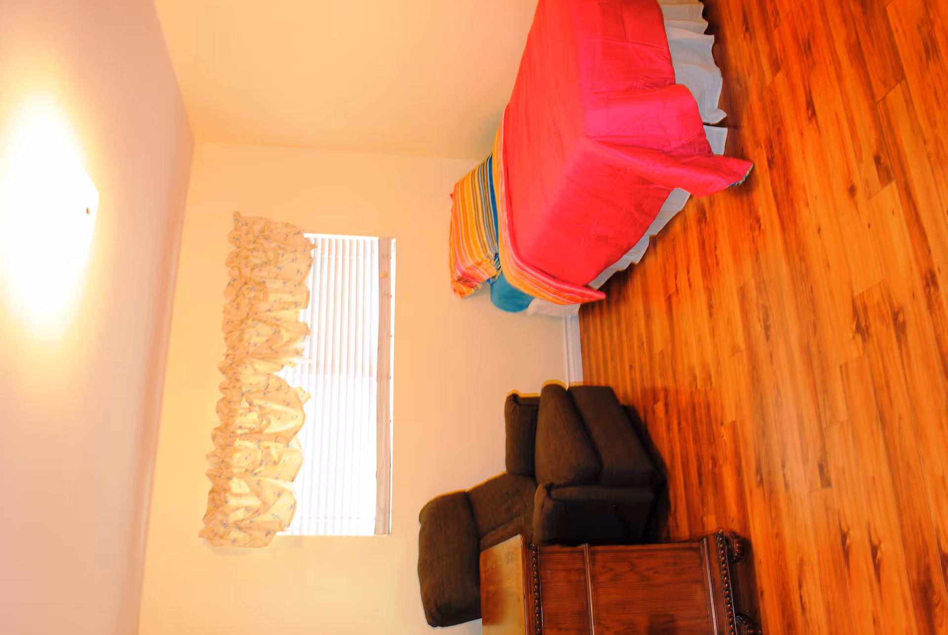 A small bedroom with a single bed covered in a bright pink bedspread and a colorful striped blanket at the foot. There is a dark brown recliner chair next to a wooden chest. The room has light-colored walls, a window with white blinds and a floral valance, and wood flooring.