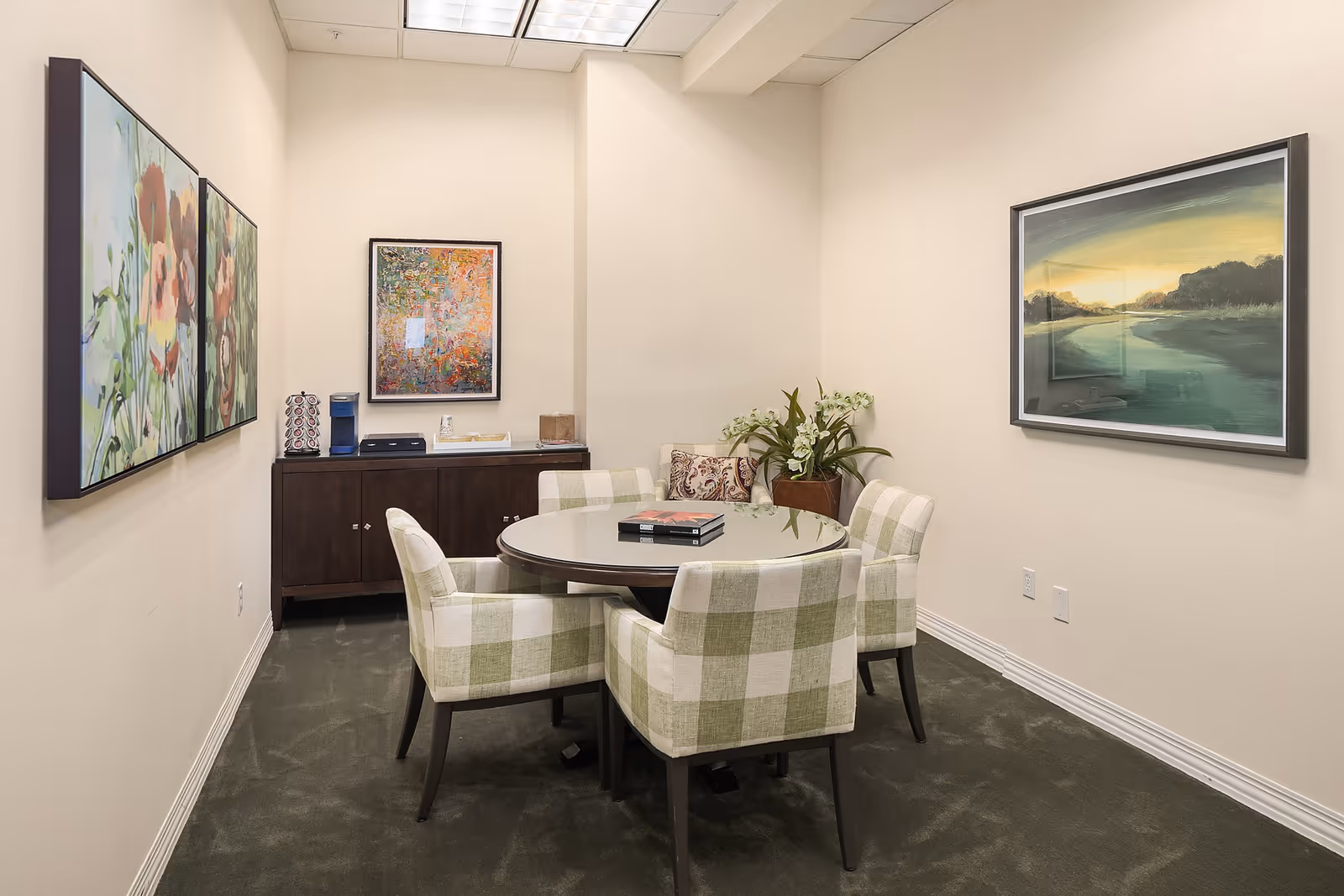 A small meeting or dining room with a round table surrounded by five green and white checkered upholstered chairs. On the table are two books. The room has beige walls adorned with three framed paintings: two floral paintings on the left wall and a landscape painting on the right wall. At the back of the room is a dark wooden cabinet with a coffee maker, a decorative container, and some small items on top. A potted plant with white flowers is placed near the cabinet.