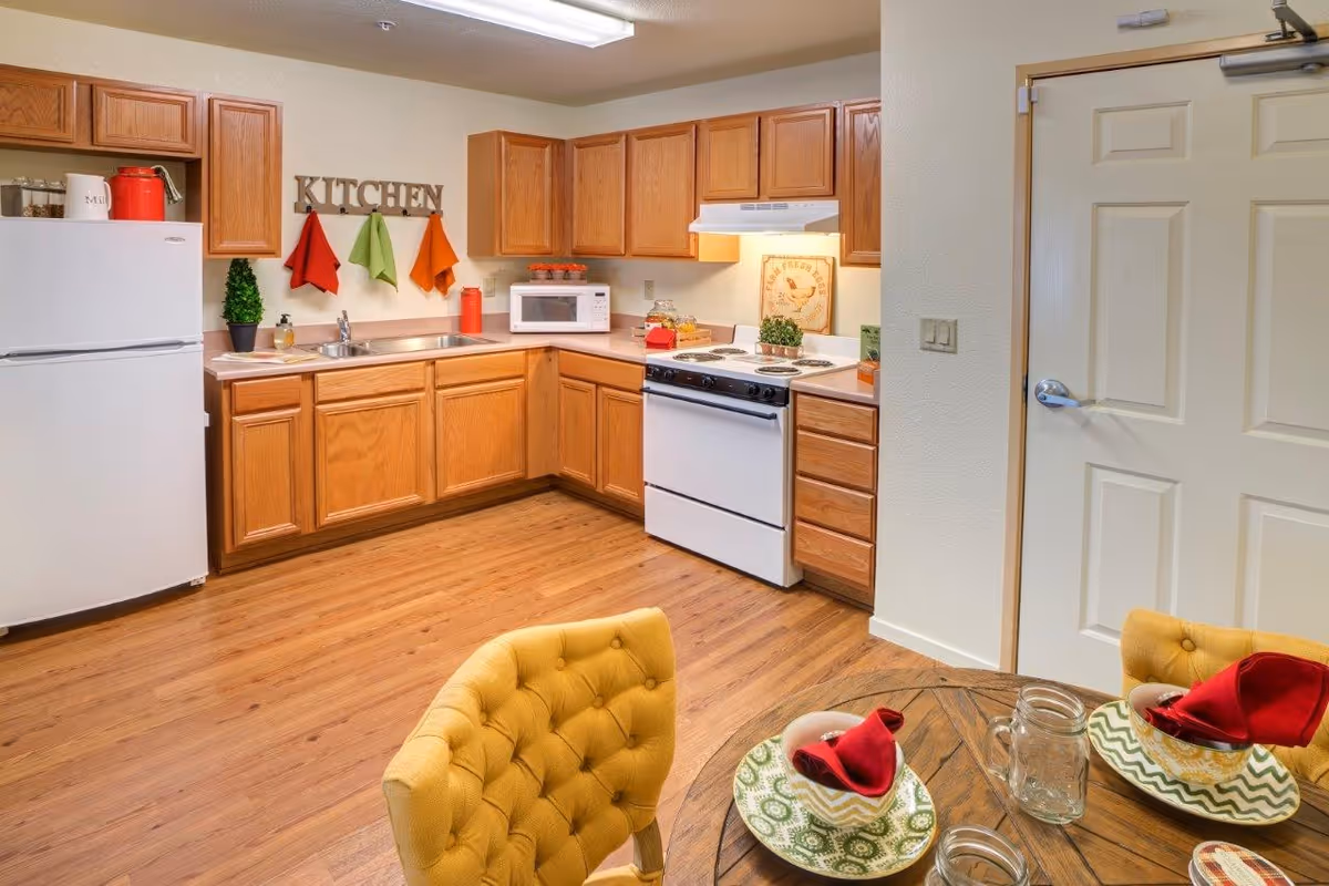 A cozy kitchen with wooden cabinets, a white refrigerator, stove, and microwave. Colorful towels hang on the wall beneath a decorative sign that reads 'KITCHEN'. In the foreground, a round dining table is set with plates, napkins, and glasses, accompanied by yellow upholstered chairs. The floor is wood, and a closed door is visible on the right side.