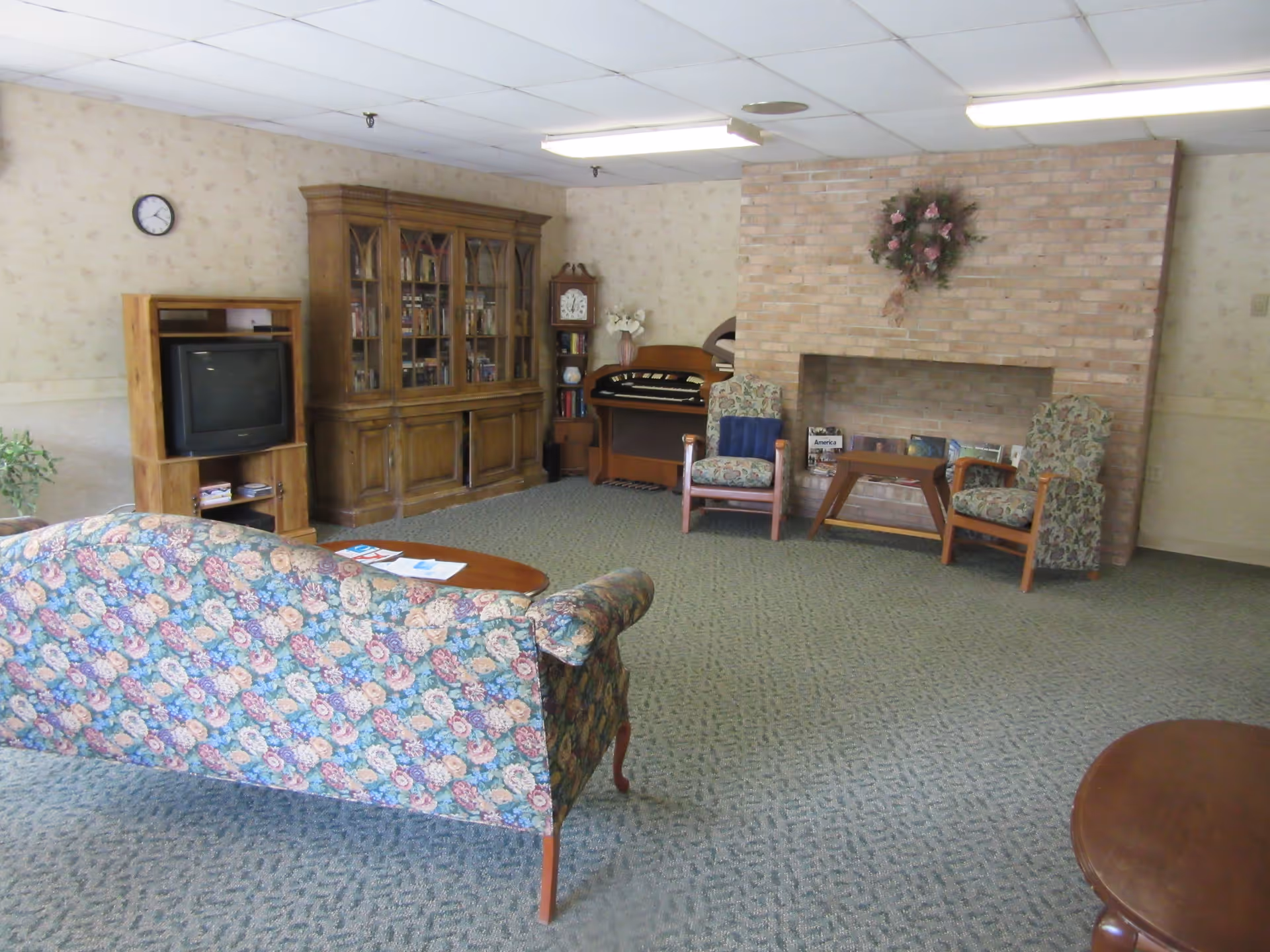 A cozy living room with floral patterned sofa and two armchairs facing a brick fireplace decorated with a floral wreath. There is a wooden coffee table in front of the sofa, a wooden cabinet with a TV, a bookshelf, and a vintage clock. The room has carpeted flooring and a drop ceiling with fluorescent lighting.
