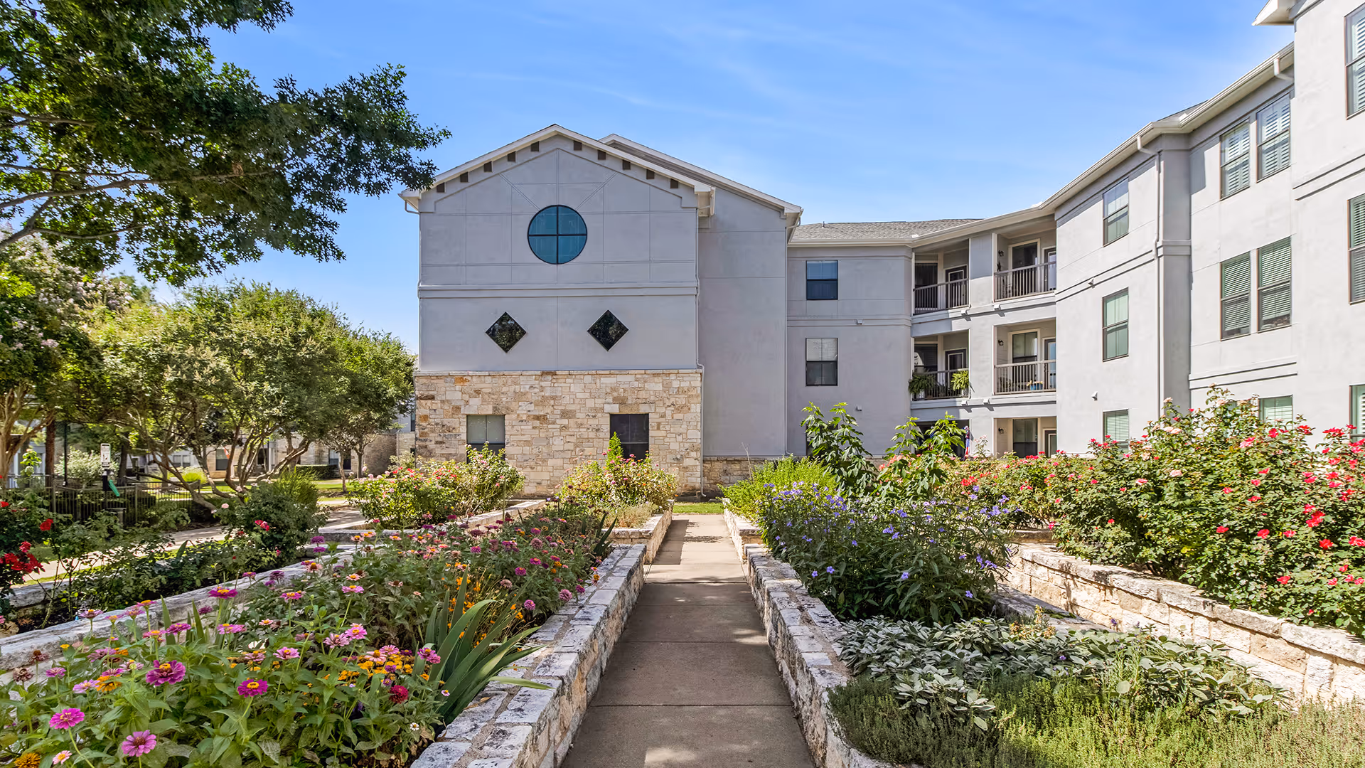 Outdoor garden area with colorful flowers and plants bordered by stone walls, leading to a multi-story building with balconies and various windows under a clear blue sky.