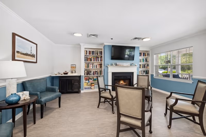 A cozy common area with a fireplace, a wall-mounted TV above it, built-in bookshelves filled with books on either side, several chairs arranged around a small table, a blue armchair next to a side table with a lamp, and a large window letting in natural light.