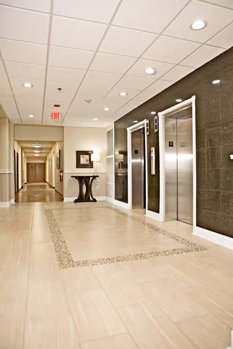 A clean and well-lit hallway in a senior living facility with two stainless steel elevators on the right side, a decorative table with a lamp and framed artwork on the wall, beige tiled floors with a patterned border, and a long corridor extending into the distance with multiple doors and ceiling lights.
