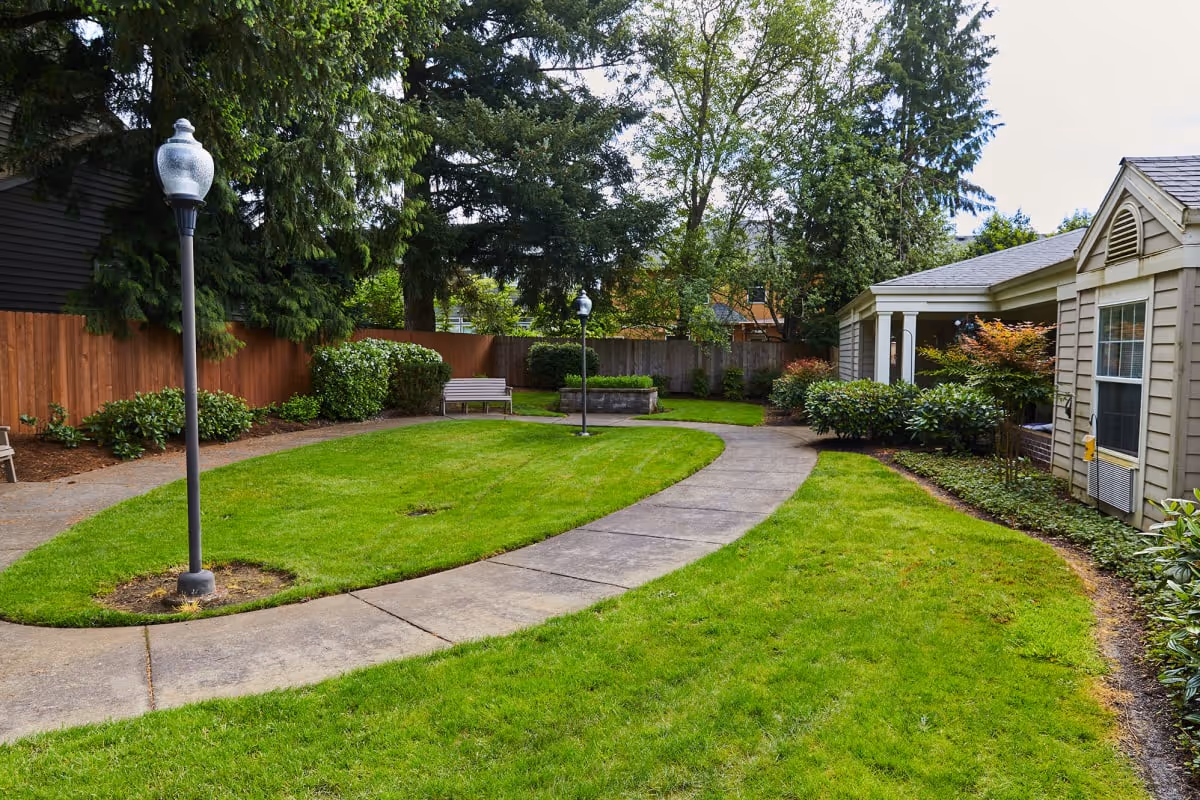 A well-maintained outdoor garden area at Jurgens Park Senior Living featuring a curved concrete walkway, green grass, bushes, trees, two lamp posts, a wooden bench, and part of a building with beige siding and a window.