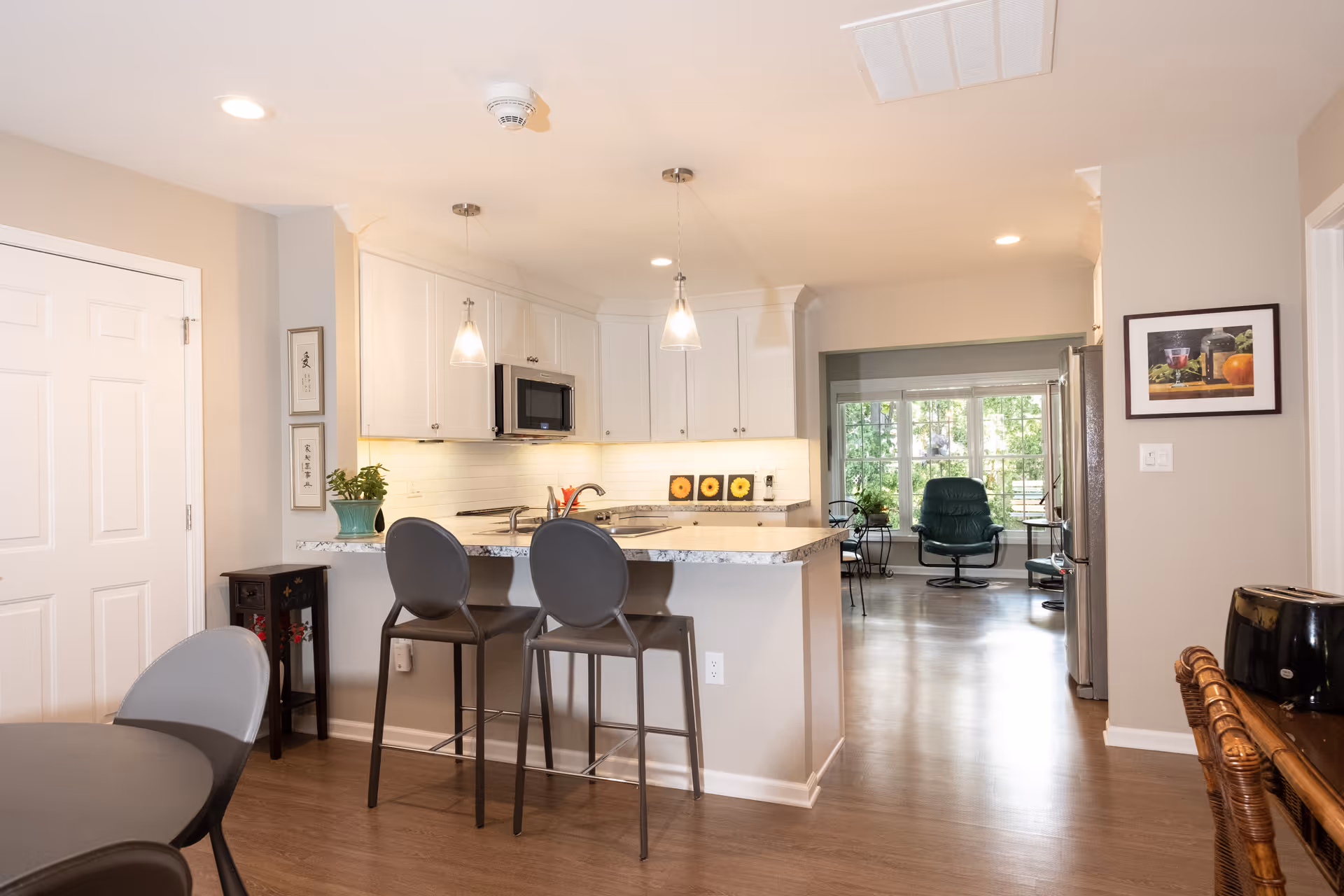 A modern kitchen area with white cabinets, a granite countertop island with two gray bar stools, and pendant lights hanging above. The kitchen opens into a living area with large windows and a green armchair. The room has light-colored walls and wooden flooring.