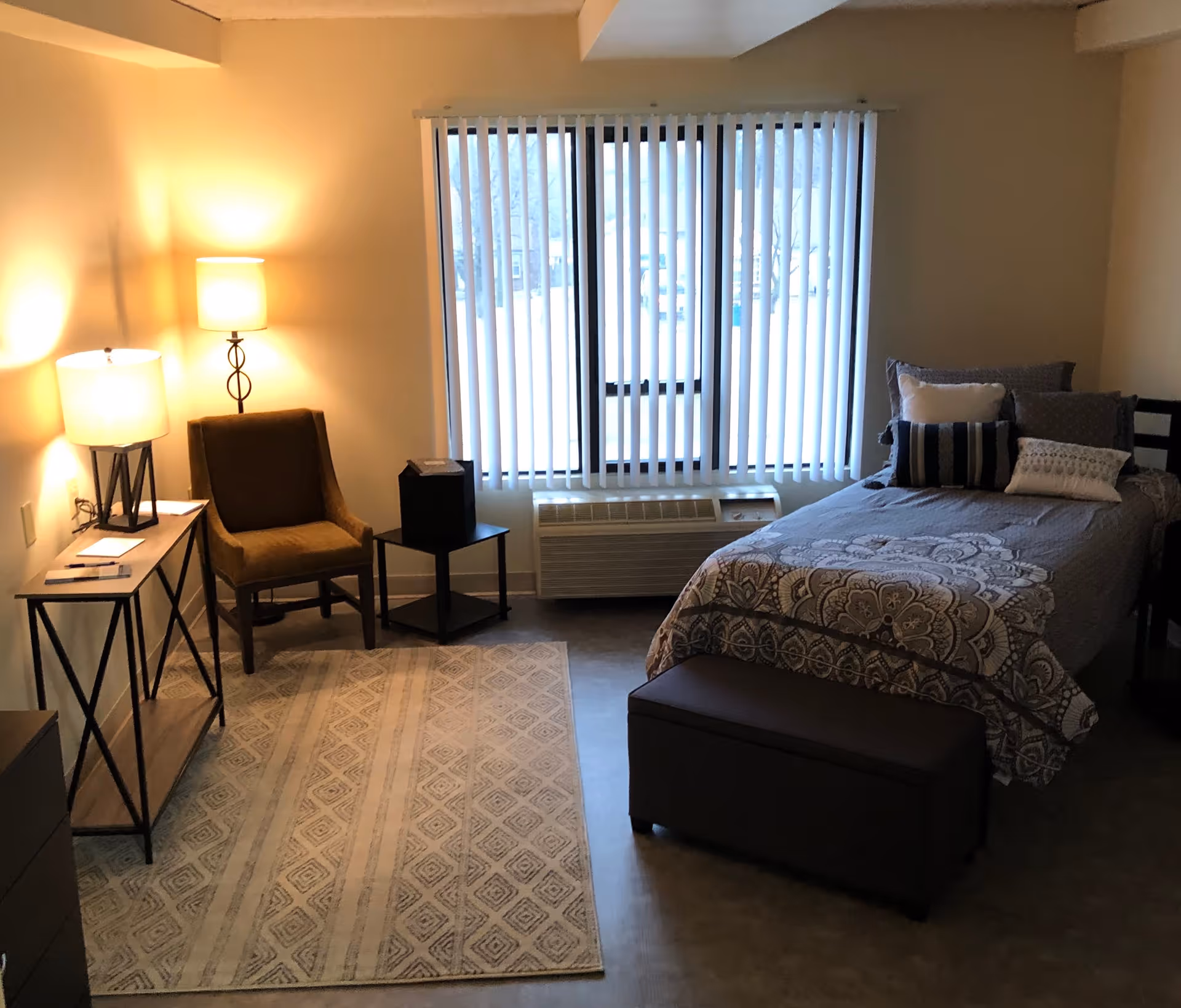 A cozy bedroom with a single bed covered in patterned bedding and several pillows. Next to the bed is a dark ottoman. A window with vertical blinds is centered on the back wall above an air conditioning unit. To the left, there is a brown upholstered chair, a small black side table, and a console table with a lamp and some items on it. The floor has a patterned area rug and the room is softly lit by two lamps.