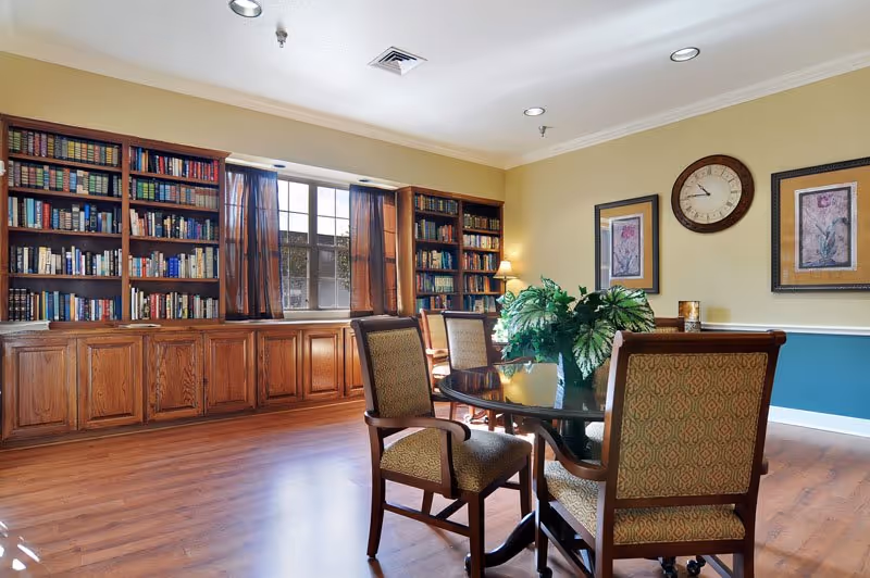 A cozy senior living common area with wooden bookshelves filled with books along the wall, a window with brown curtains letting in natural light, a round glass table with four upholstered chairs, a green potted plant centerpiece on the table, a wall clock, and framed floral artwork on the walls.